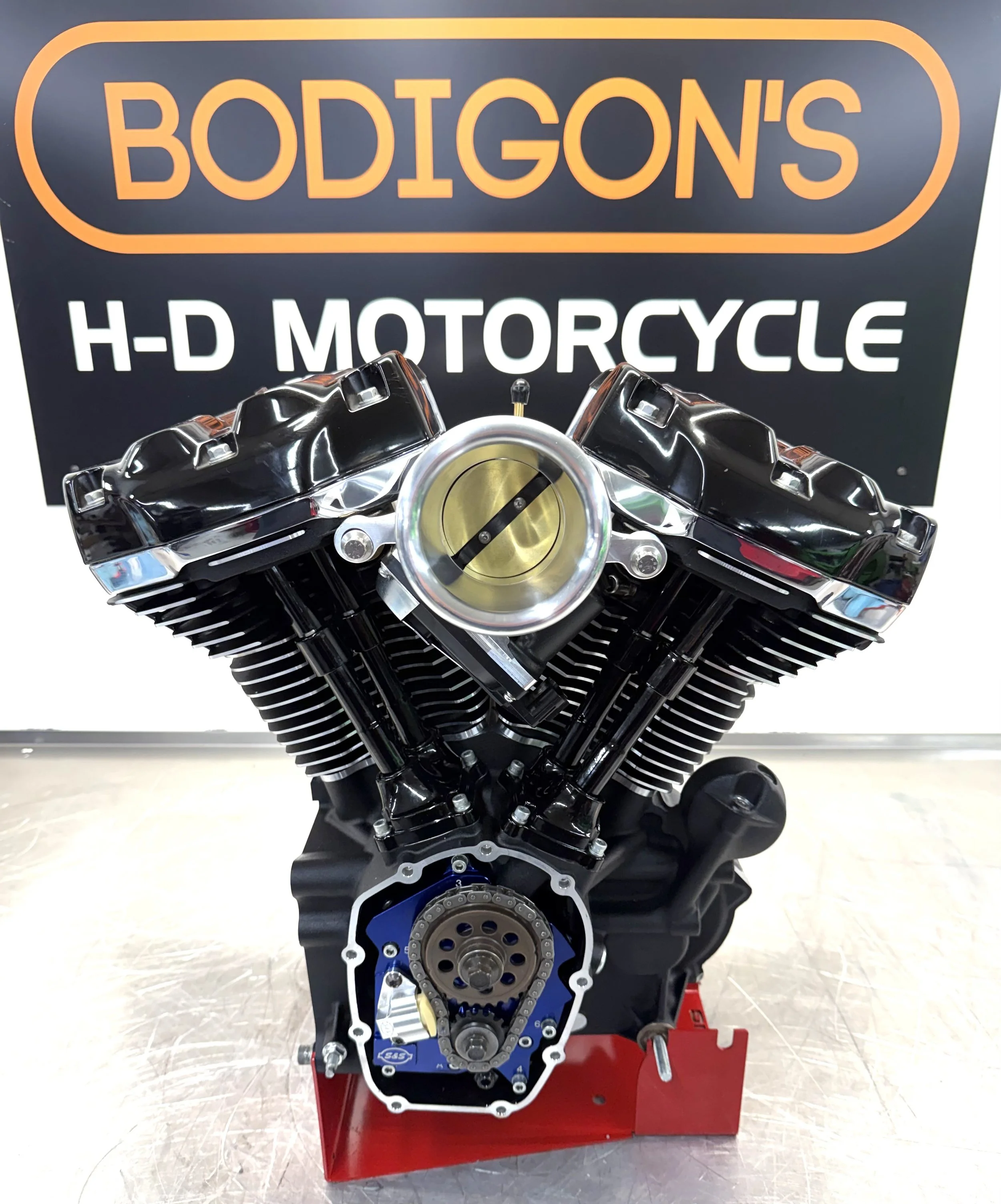 An internal combustion motorcycle engine on a red engine stand in front of a sign that reads 'Bodigon's H-D Motorcycle'.
