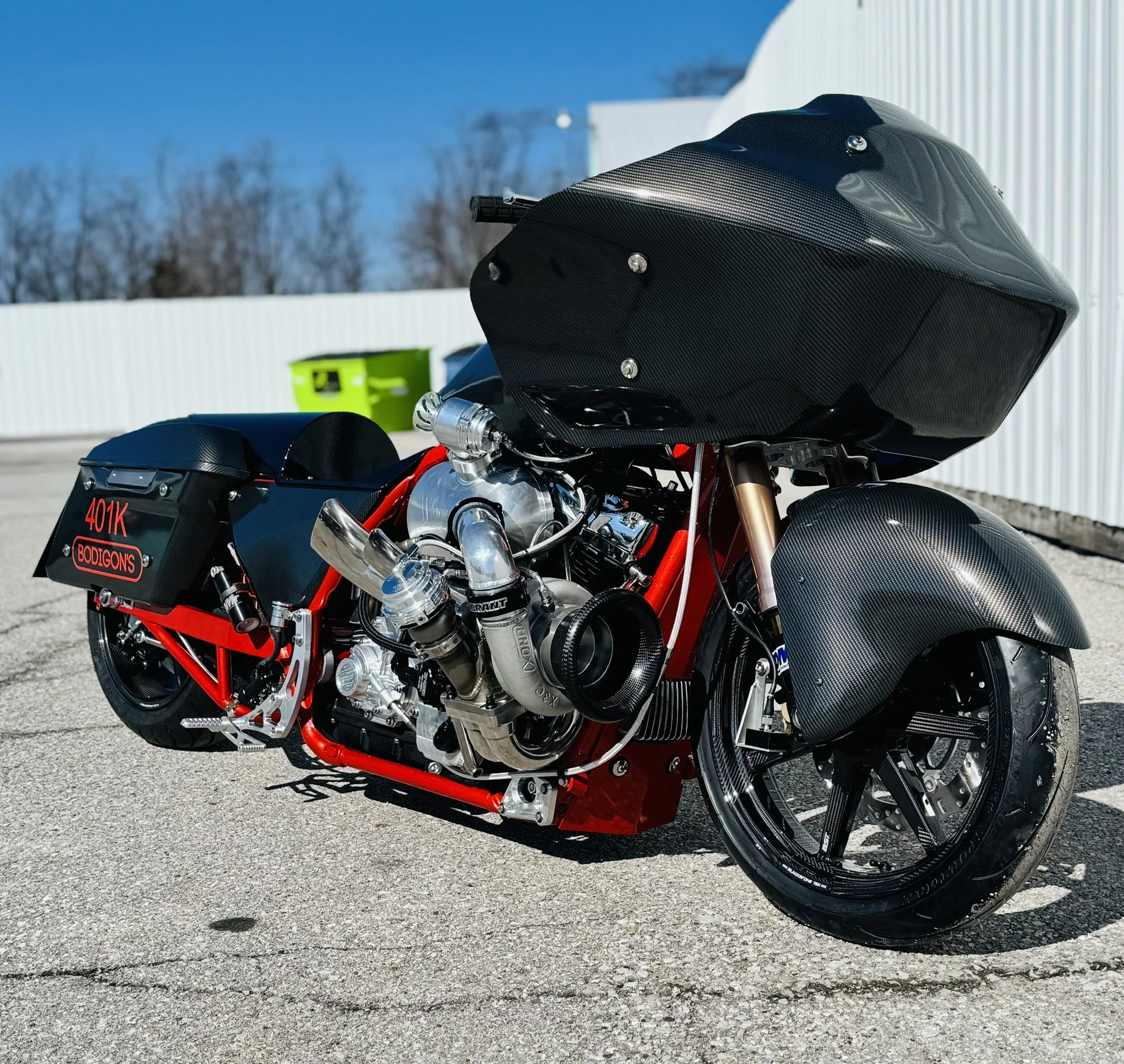 Close-up of a black and red sport motorcycle with a textured black fairing and exposed engine components, parked on a paved surface with a white building and leafless trees in the background under a clear blue sky.