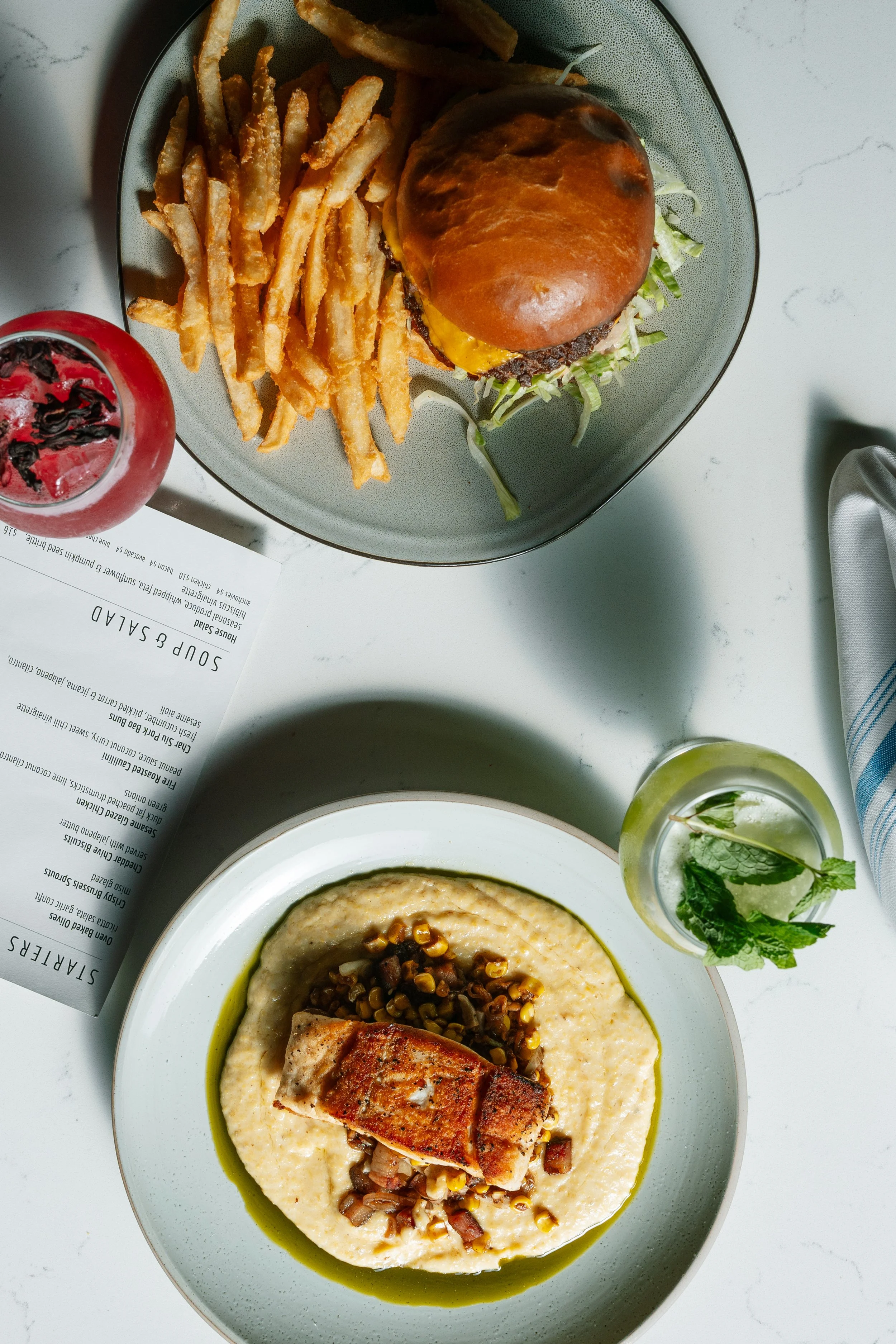 A plate of food featuring a piece of grilled fish with a topping of corn and black beans on a bed of creamy mashed potatoes. There is a green sauce around the mashed potatoes. In the background, there is a side dish of fries and a burger with lettuce, tomato, cheese, and a brioche bun. A glass of a red drink with ice and a garnish of berry and mint leaves are also visible.