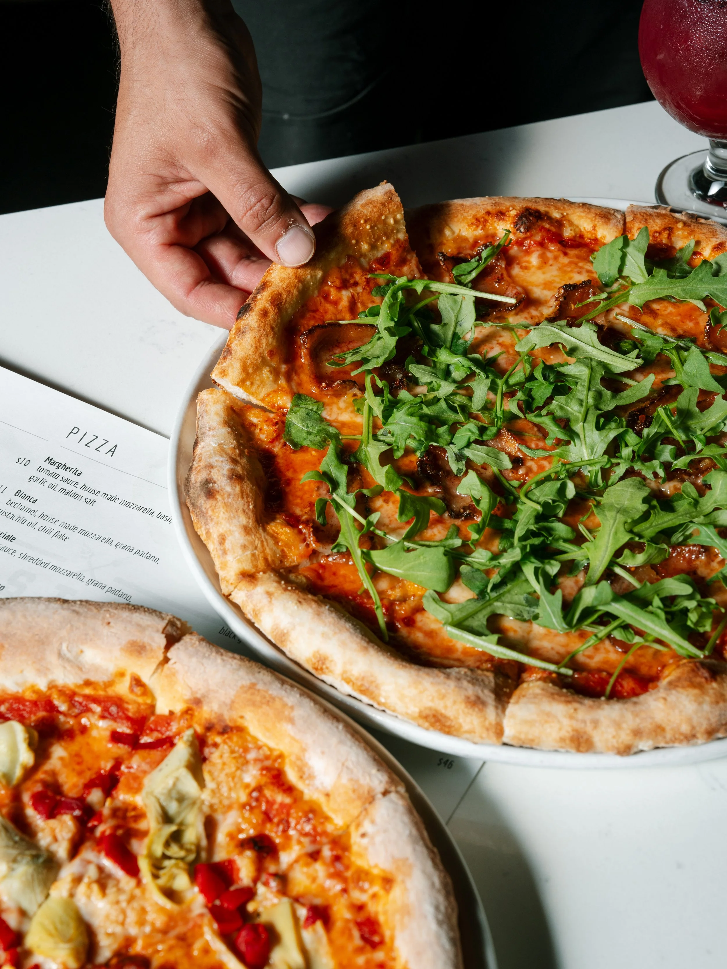 A person is holding a slice of pizza topped with basil and other herbs. There are other pizzas on the table, one with jalapeños and red pepper and another with artichokes and pimientos, along with a glass of red drink.