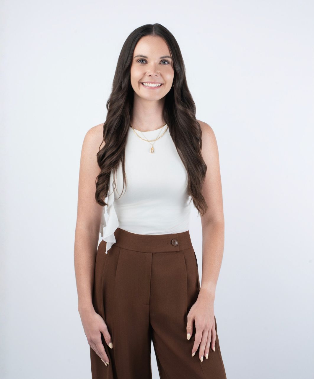 A young woman with long dark hair, wearing a white sleeveless top and brown high-waisted pants, smiling and standing against a plain white background.