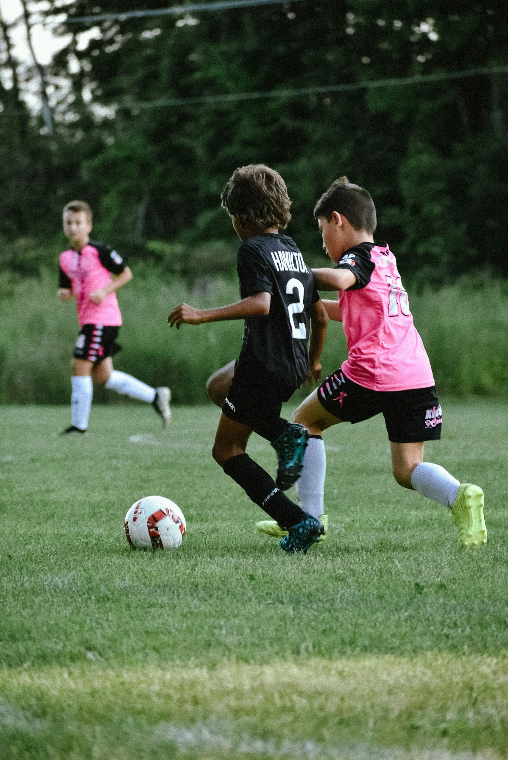 Young boys playing soccer on a grassy field, two in black uniforms and one in pink, engaged in a match during daytime.