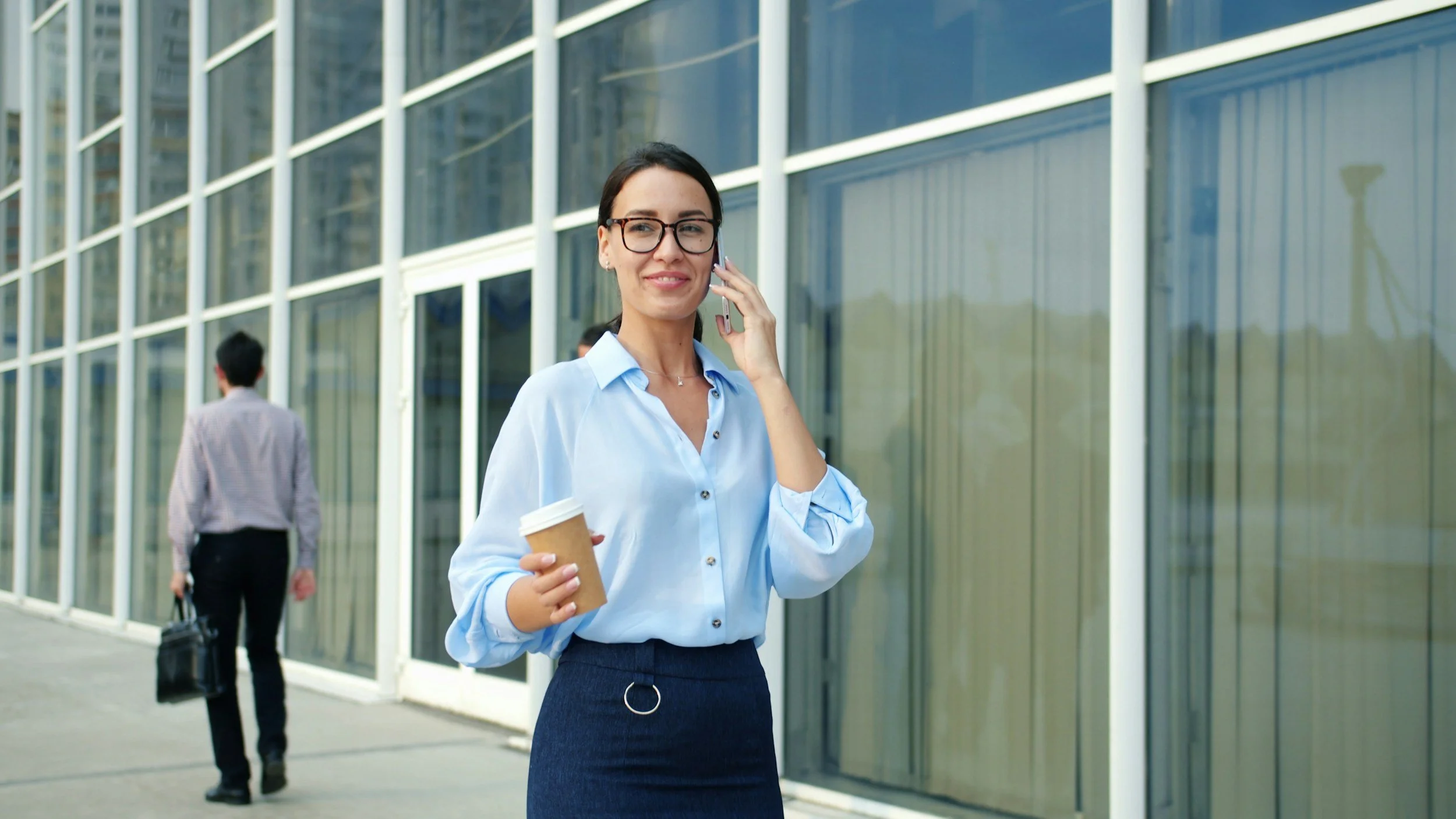 A woman with glasses in business attire, holding a coffee cup, talking on her phone outside a modern glass building.