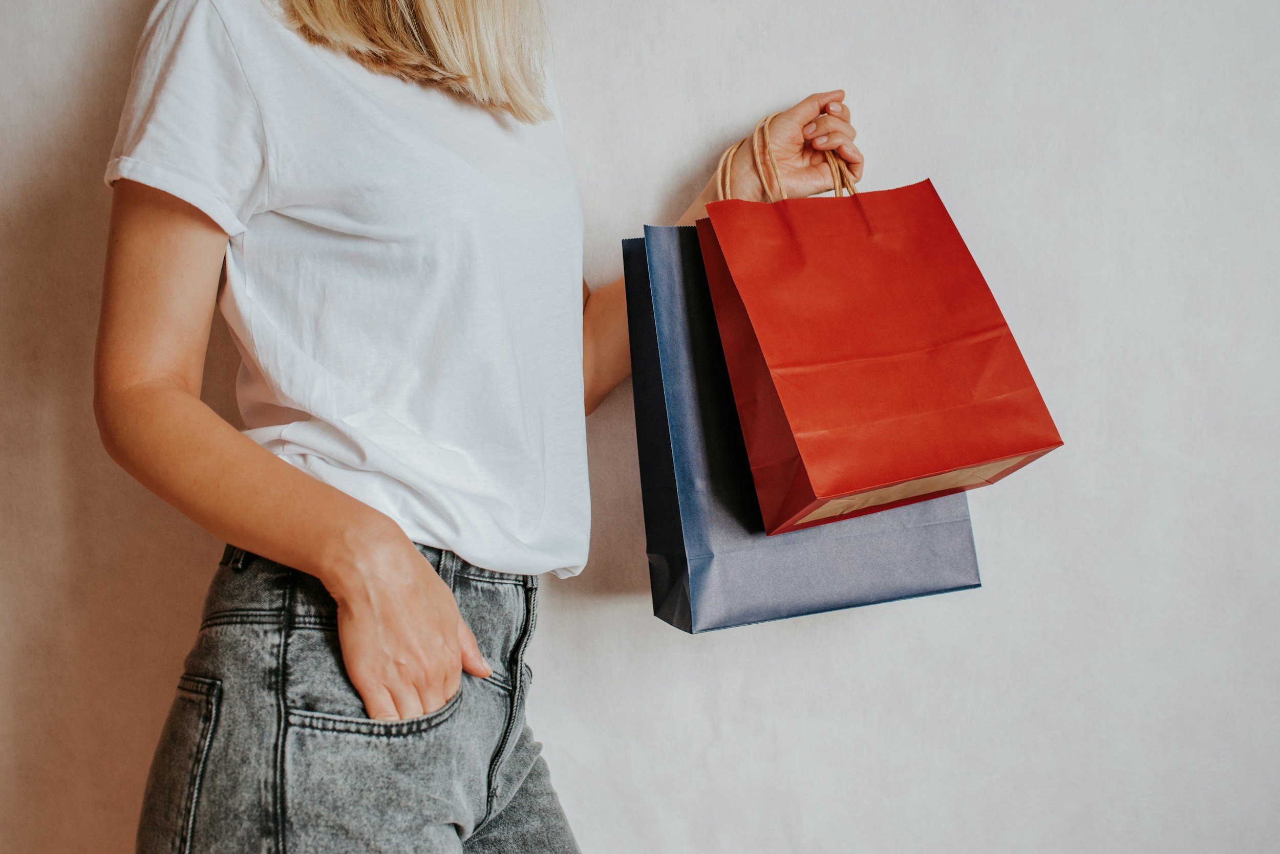 Person holding three shopping bags, wearing a white t-shirt and gray jeans, against a plain wall.