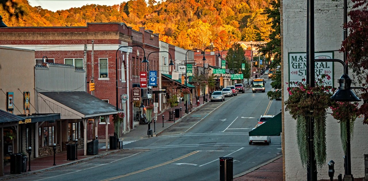 A small town street during autumn with colorful trees in the background, brick buildings, parked cars, street lamps, and signs along the sidewalk.