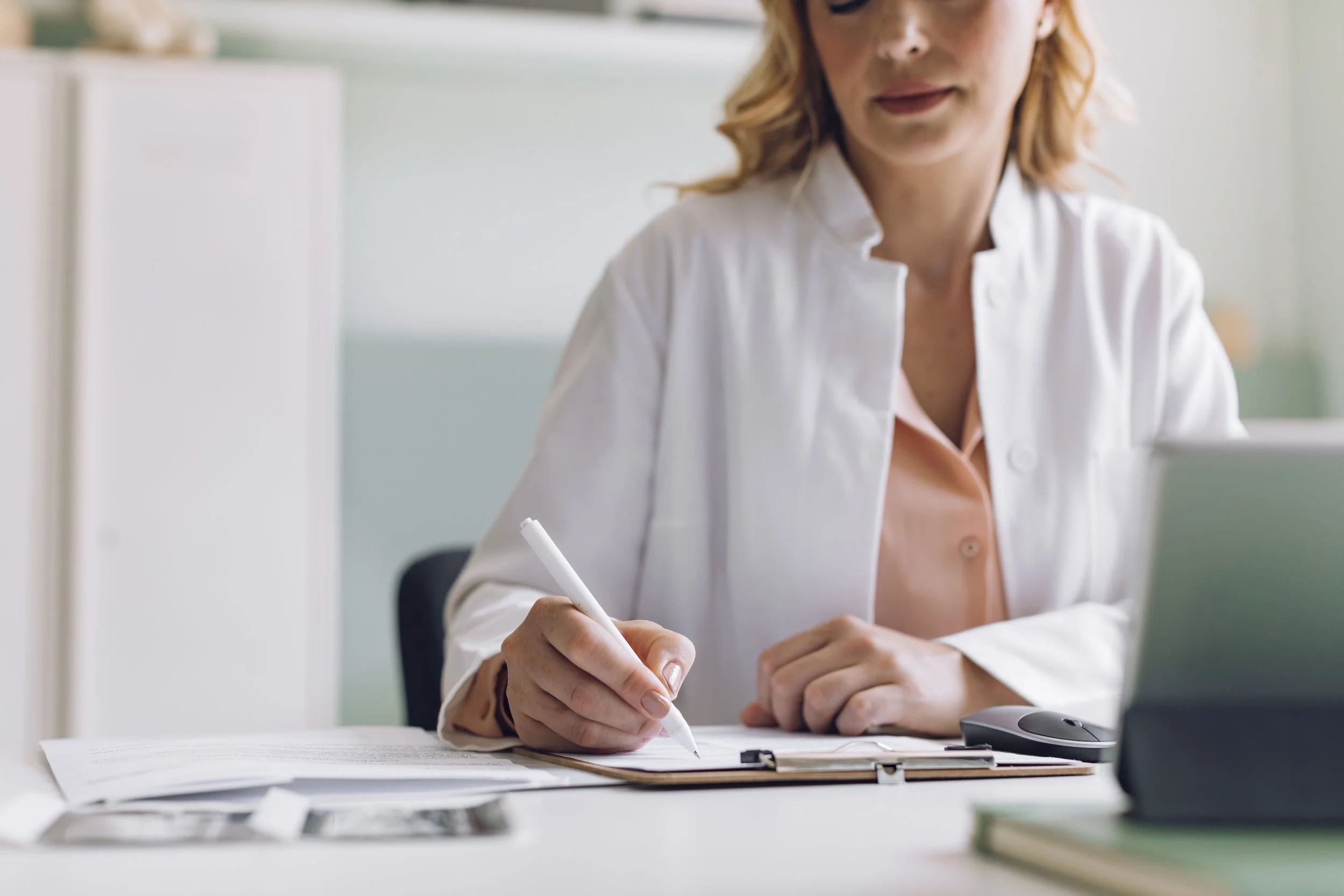 A woman in a white coat sitting at a desk, writing on a clipboard.