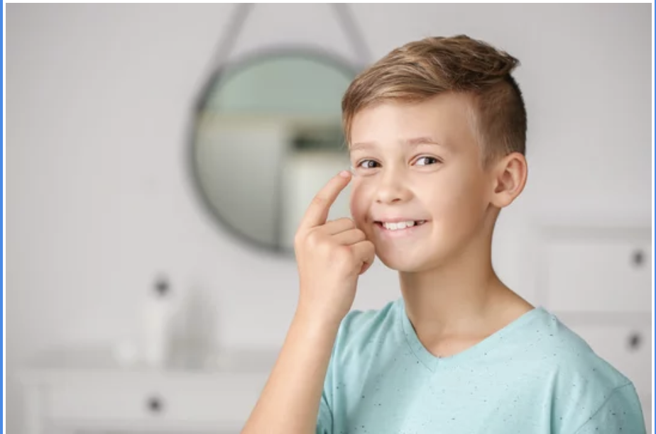 A young boy with short, light brown hair smiling and touching his face in a bathroom with a round mirror in the background.