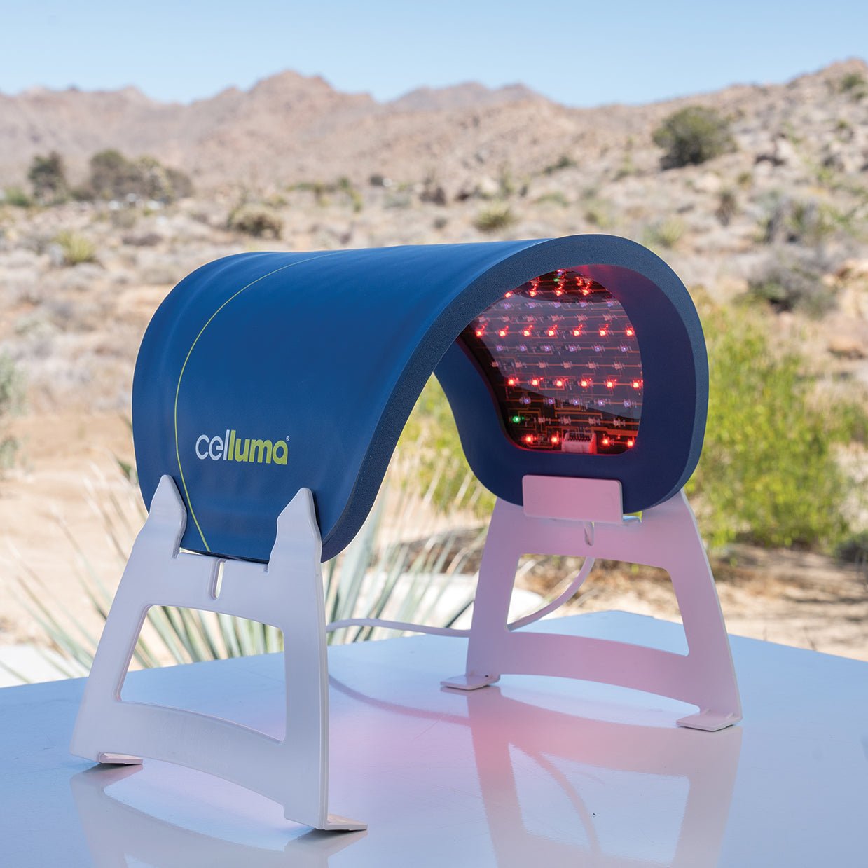 A portable medical device labeled 'celluma' with a curved blue housing and red LED lights inside, mounted on a white stand in an outdoor desert setting with mountains and cacti in the background.