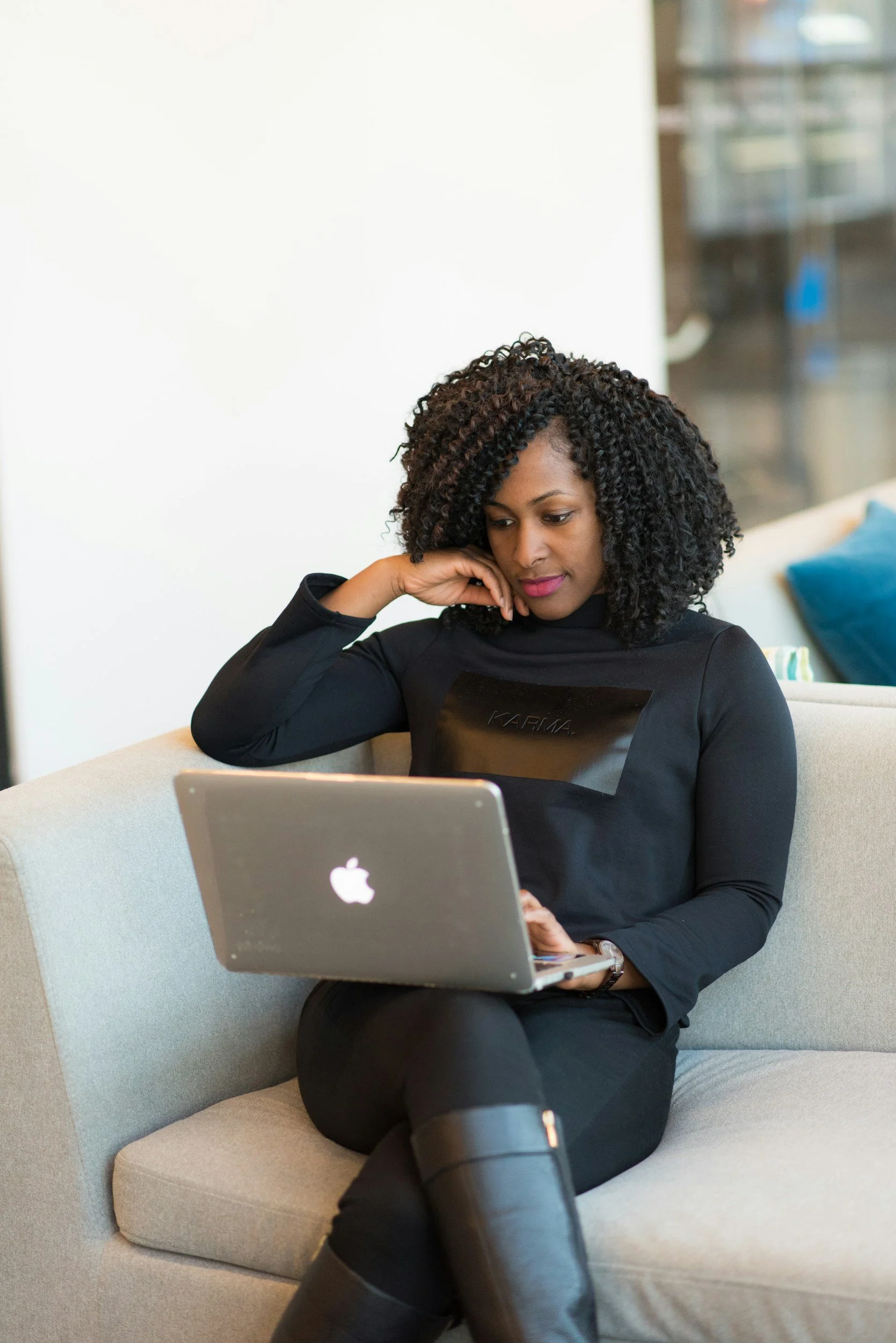 A woman sitting on a beige sofa, working on a silver MacBook, with her head resting on her left hand, in a modern indoor space.