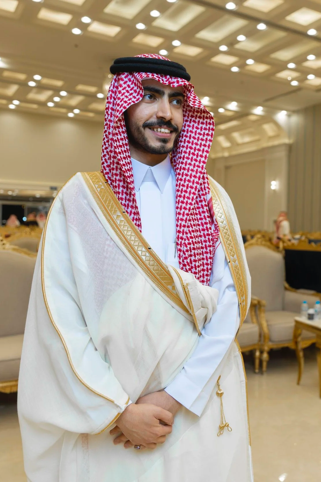 A man in traditional Middle Eastern attire, including a white thobe, a gold-embroidered bisht, and a red and white checkered keffiyeh, standing indoors with a smile.