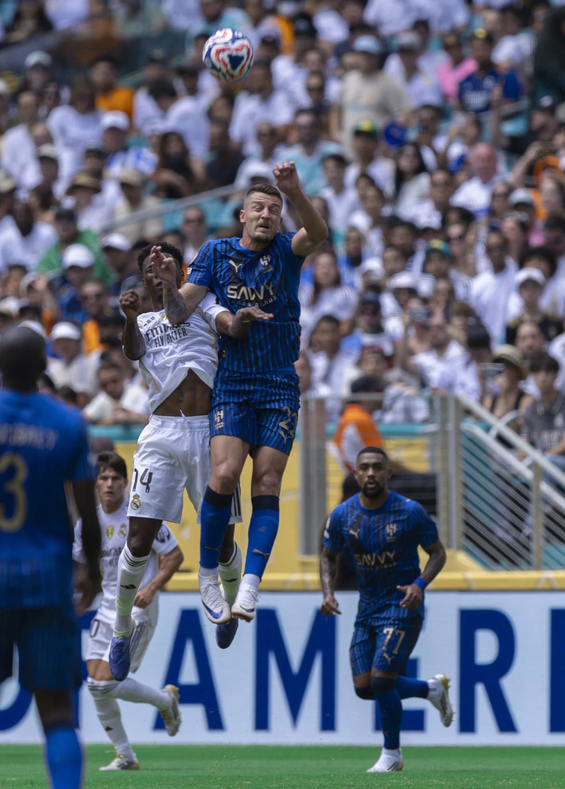 Two soccer players from opposing teams jump to head the ball during a game, with a large crowd of spectators in the background.