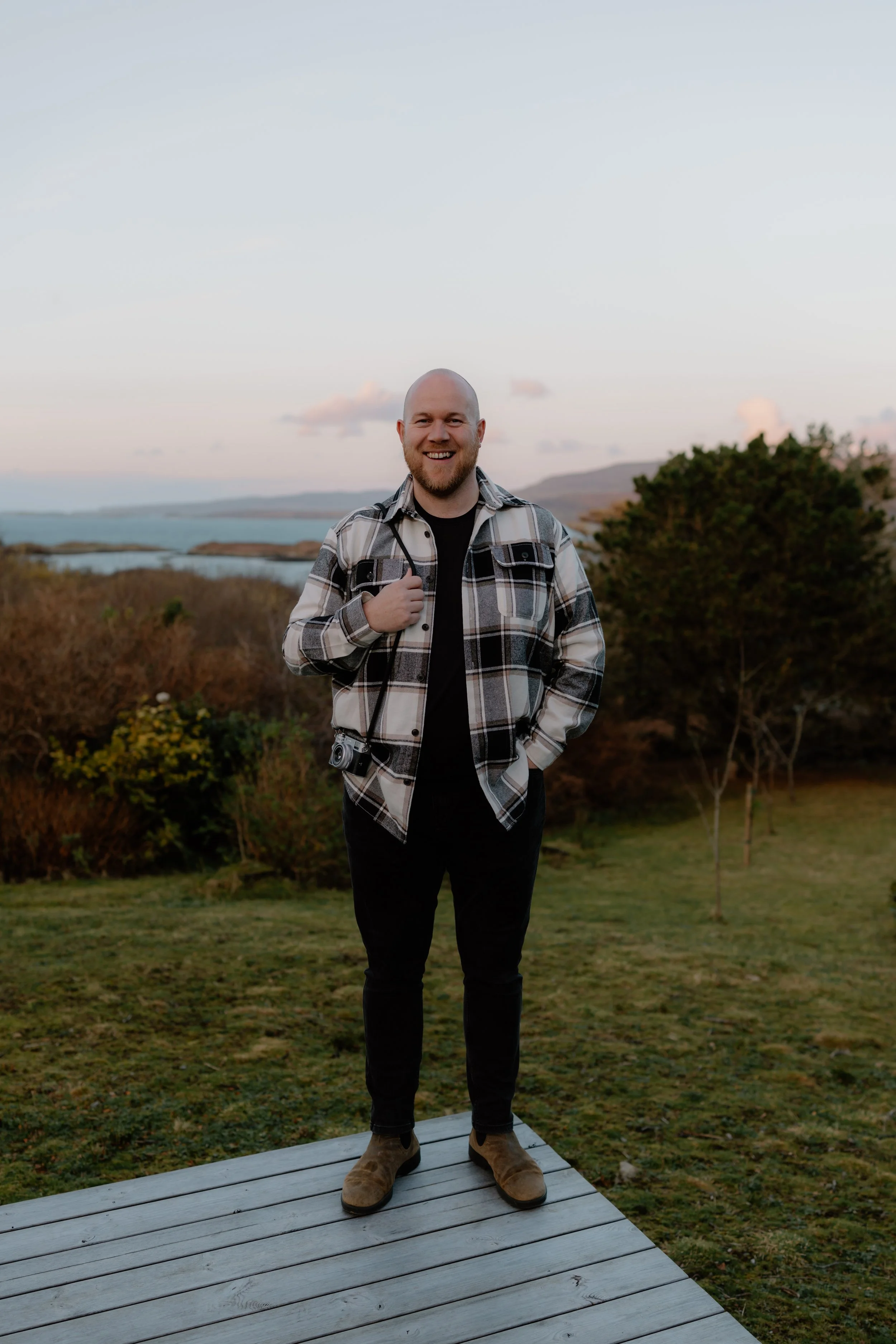 A smiling man with a beard and a bald head, wearing a plaid shirt, black pants, and brown shoes, standing on a wooden platform outdoors during sunset, with a landscape of water, hills, and bushes in the background.