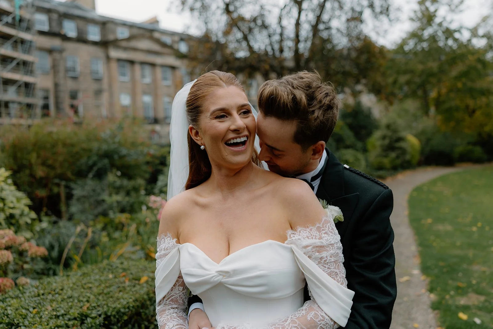 A bride in a white wedding dress and a groom in a black suit smiling and embracing outdoors in a garden in Edinburgh New Town with a building in the background.