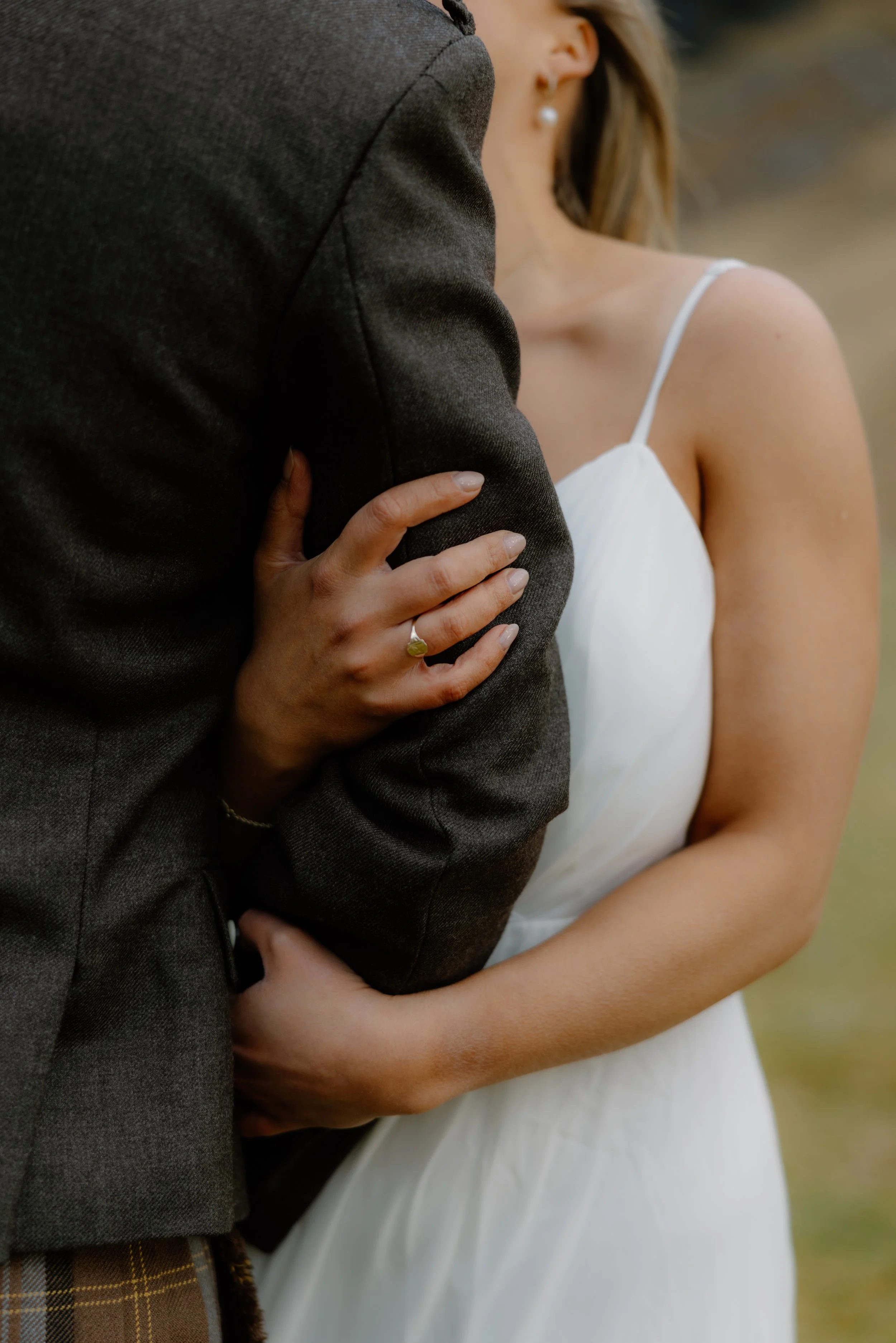 A woman in a white dress with an off-shoulder design embraces someone, with her left hand resting on their arm, showing a gold ring with a green gemstone on her ring finger.