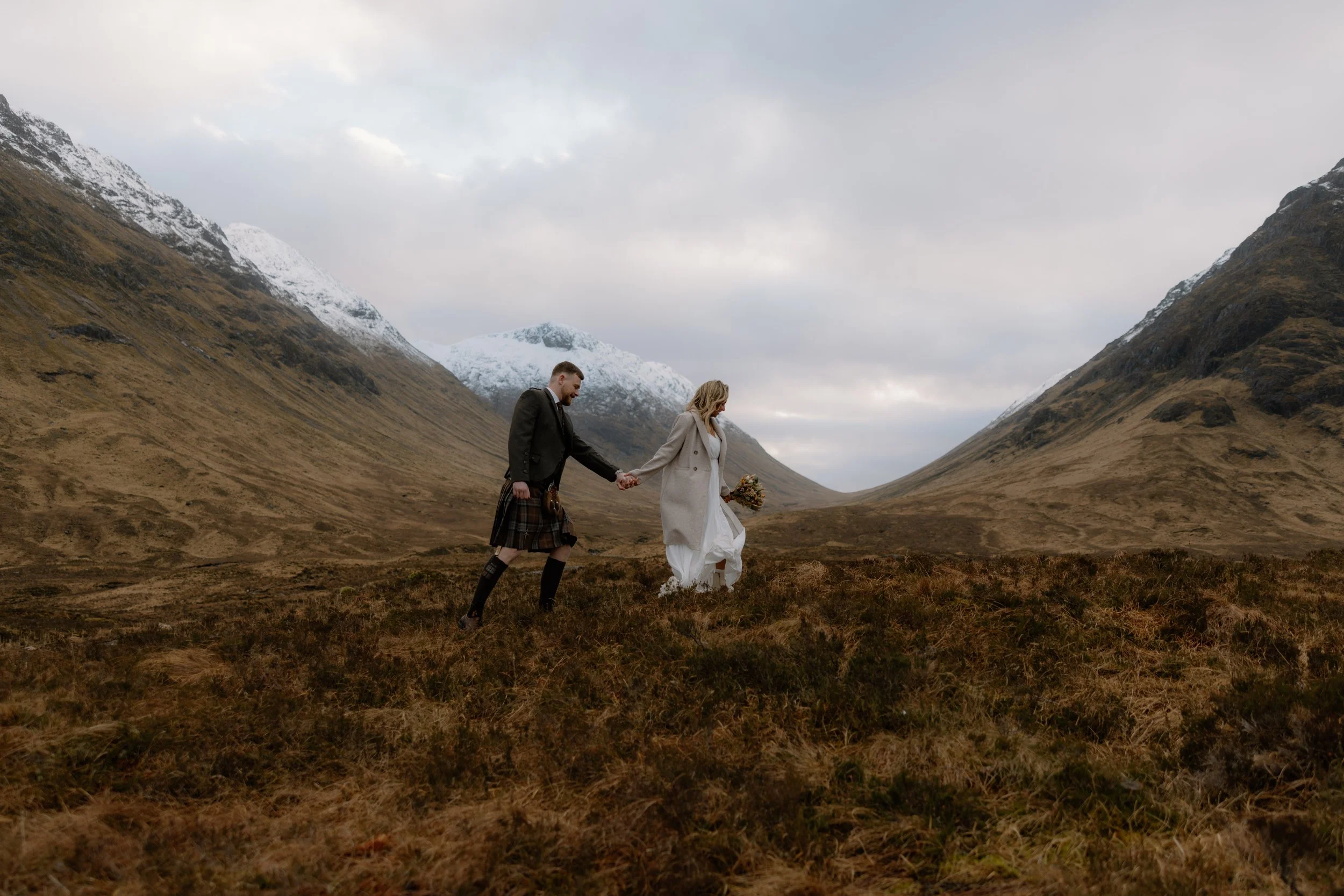 A couple holding hands walks through a valley with snow-capped mountains of Glencoe in the background, forest, and overcast sky.