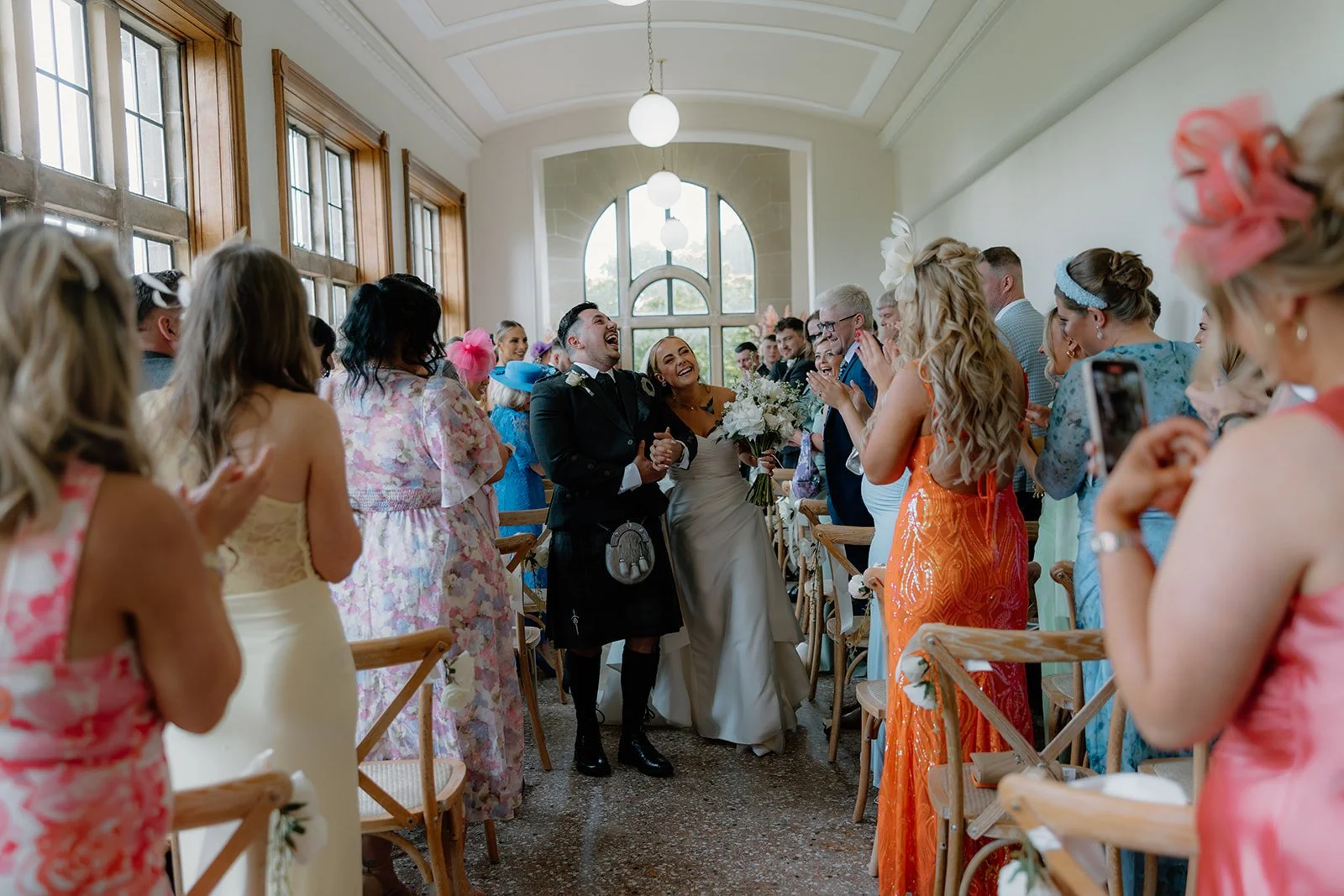 Bride and groom walking down the aisle, smiling and laughing, surrounded by guests in colourful attire inside a bright, spacious space inside Kinmount House with large windows and high ceilings.