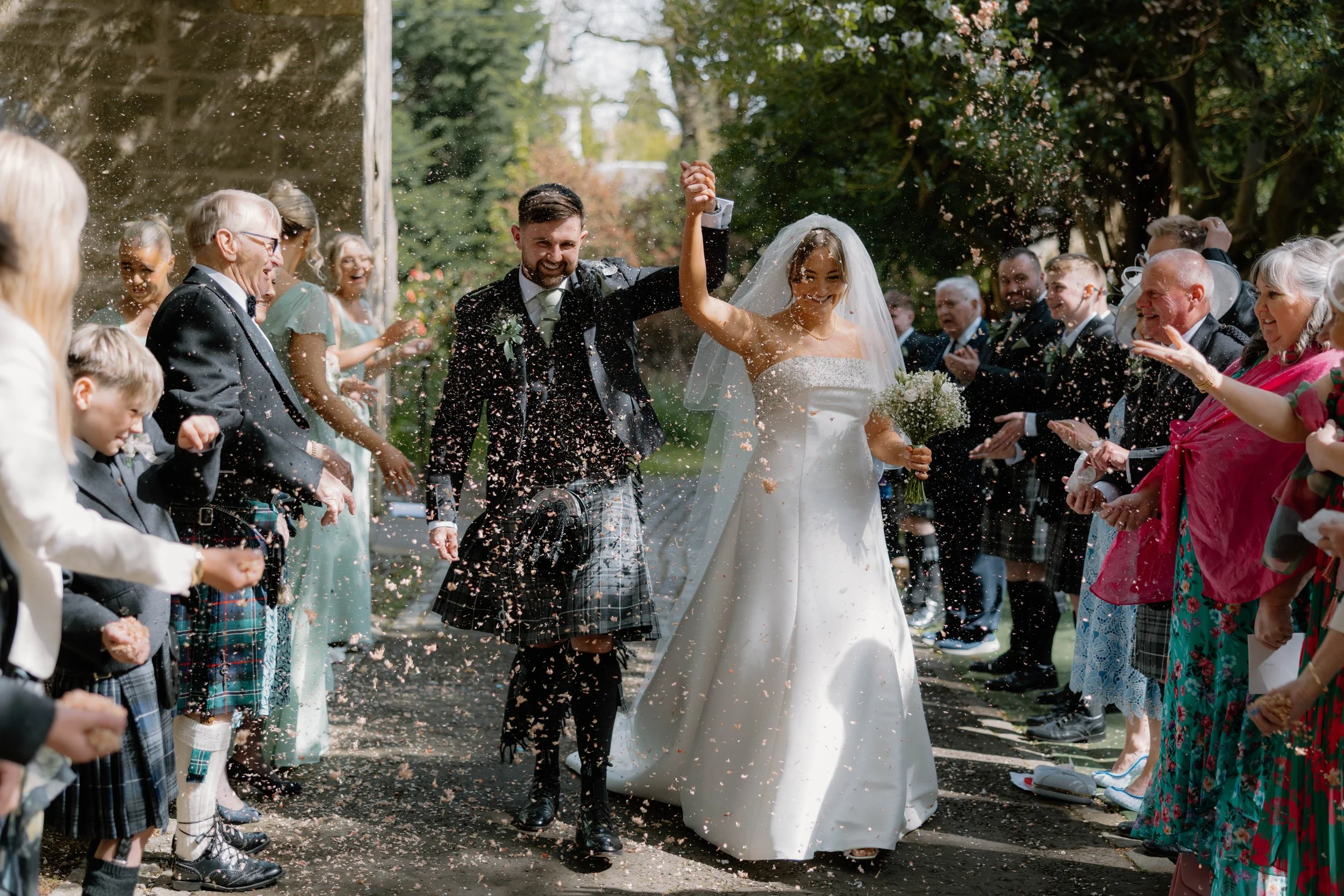 Bride and groom walking hand in hand, smiling, as they are showered with rice or confetti, surrounded by wedding guests outdoors during daytime.