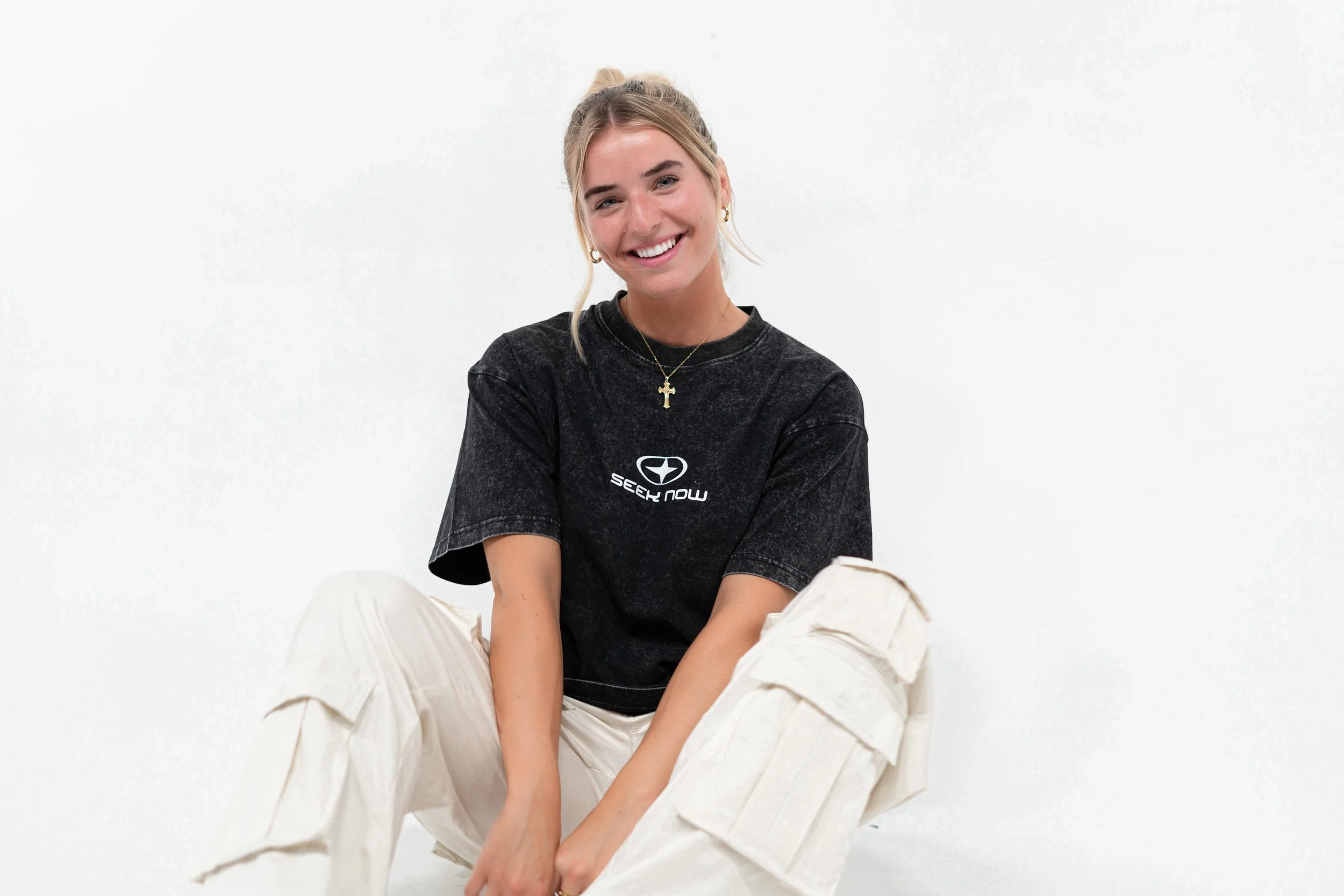 Young woman sitting on the floor, smiling, wearing a black t-shirt with a logo and the words 'Seek Now', and beige cargo pants against a plain white background.