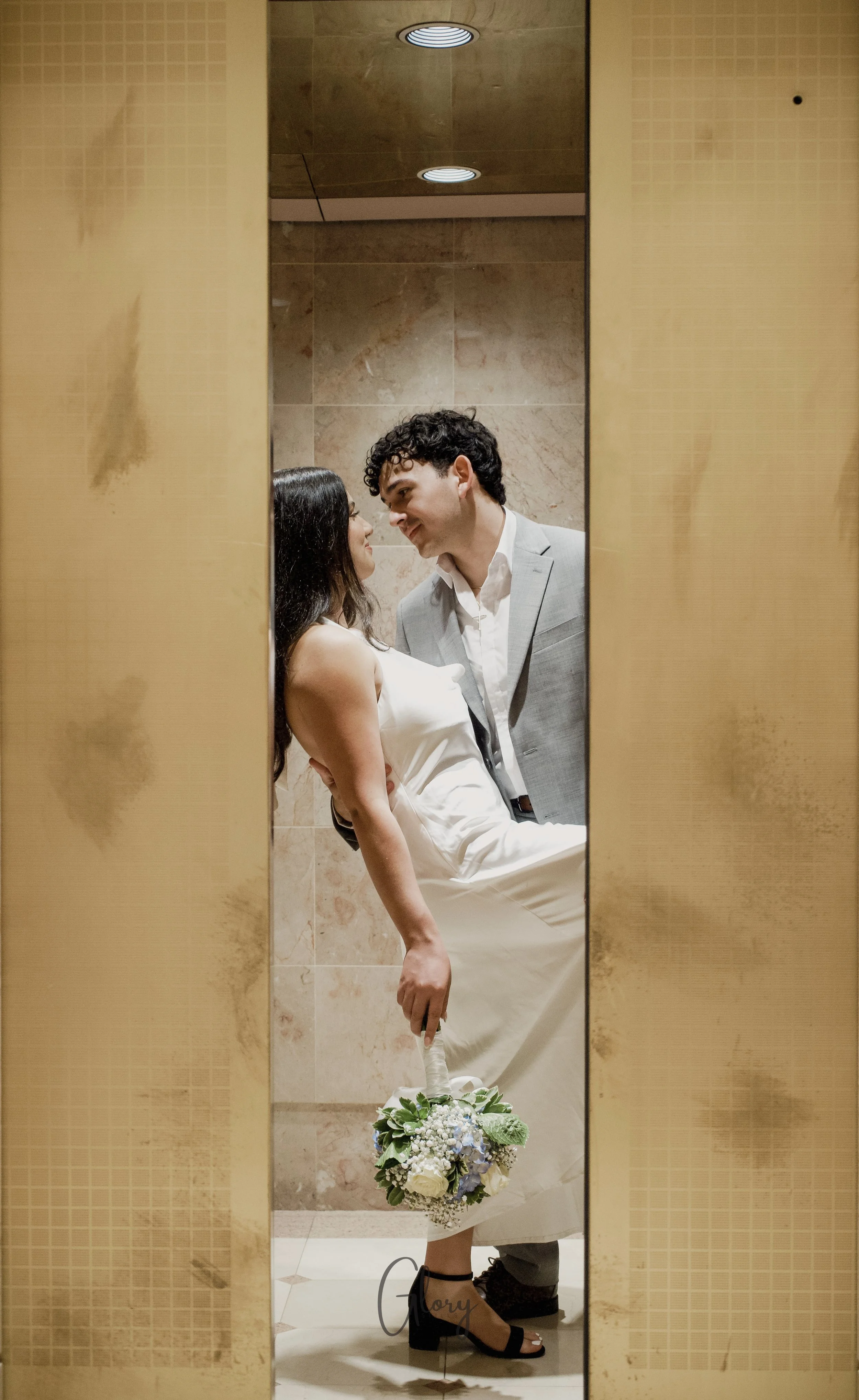 A man in a light gray suit and a woman in a white dress are sharing an intimate moment in an elevator, with their faces close together, and the woman holding a bouquet of white roses, blue flowers, and greenery, while leaning back against the elevator wall.