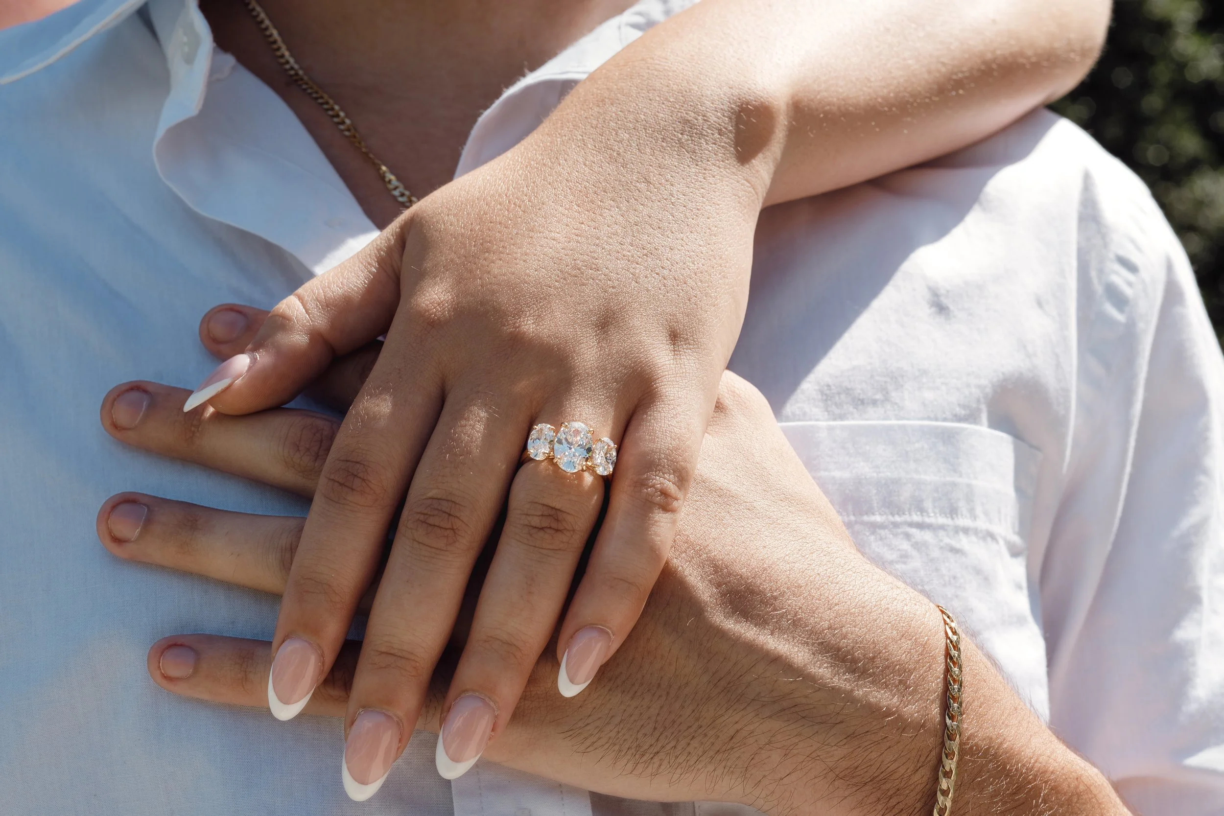 Close-up of a woman's hand with a large diamond engagement ring resting on a man's chest, both wearing white shirts, outdoors with blurred trees in background.