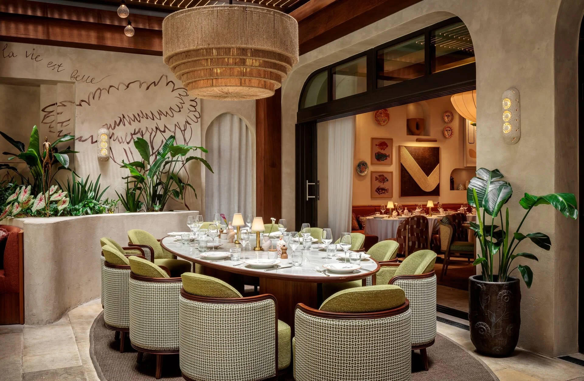 Elegant restaurant interior with a large oval dining table set with white tableware and glassware, surrounded by green upholstered chairs with wooden frames. The room features a large woven chandelier, indoor plants, and wall art on beige walls, with