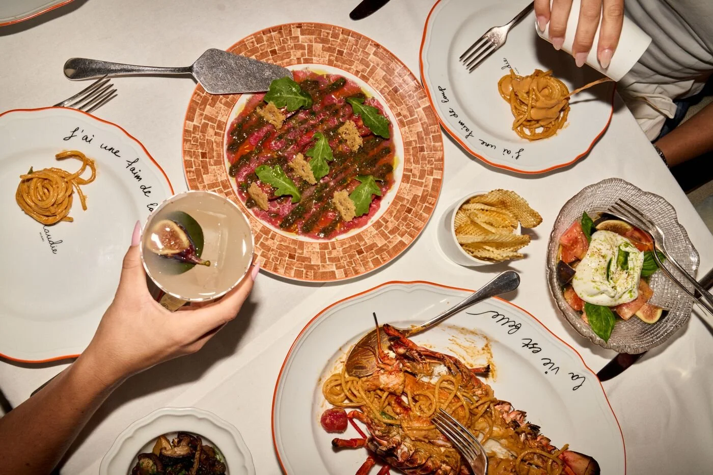 A table set with various Italian dishes including pasta, lobster, salad, and a drink, with a person's hand holding a wine glass and another person using a fork.