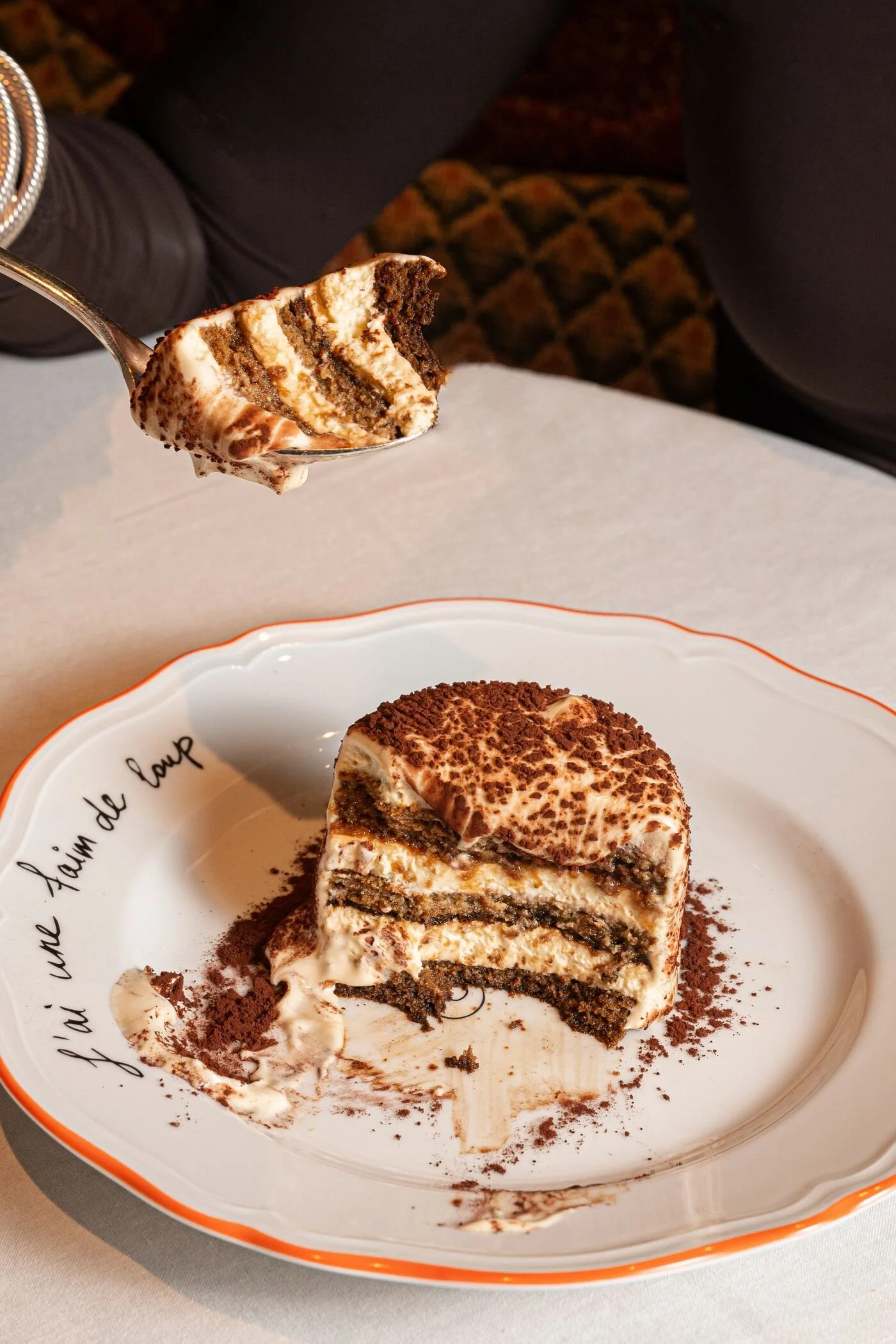 A partially eaten slice of tiramisu on a decorative plate with orange trim, surrounded by cocoa powder and cream, with a spoon holding a bite of tiramisu.