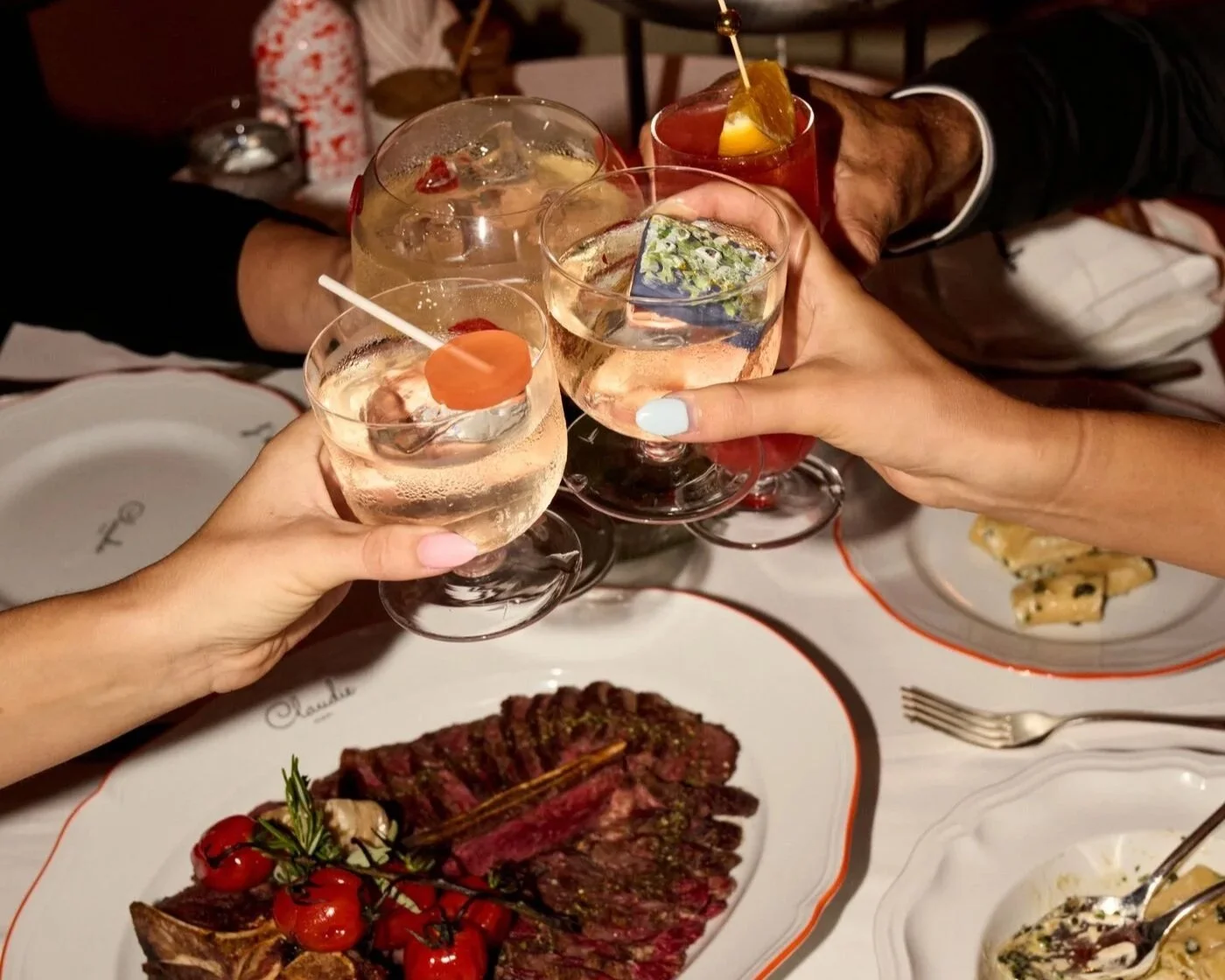 Toast with drinks and a meal on a table, including a plate of steak with cherry tomatoes and a dish of pasta.