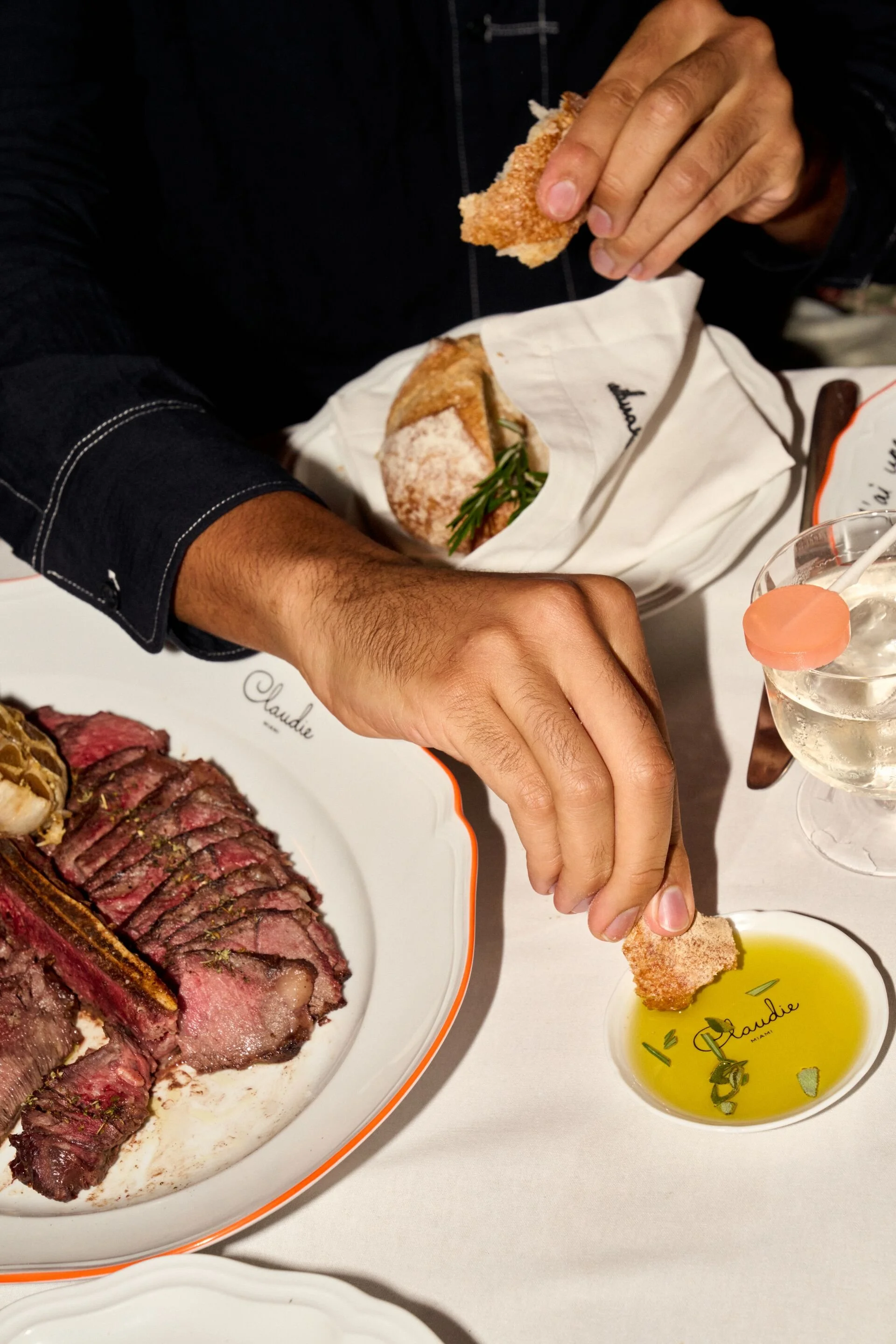 Person holding a piece of bread dipped in olive oil with herbs on a table with sliced steak and roast beef.