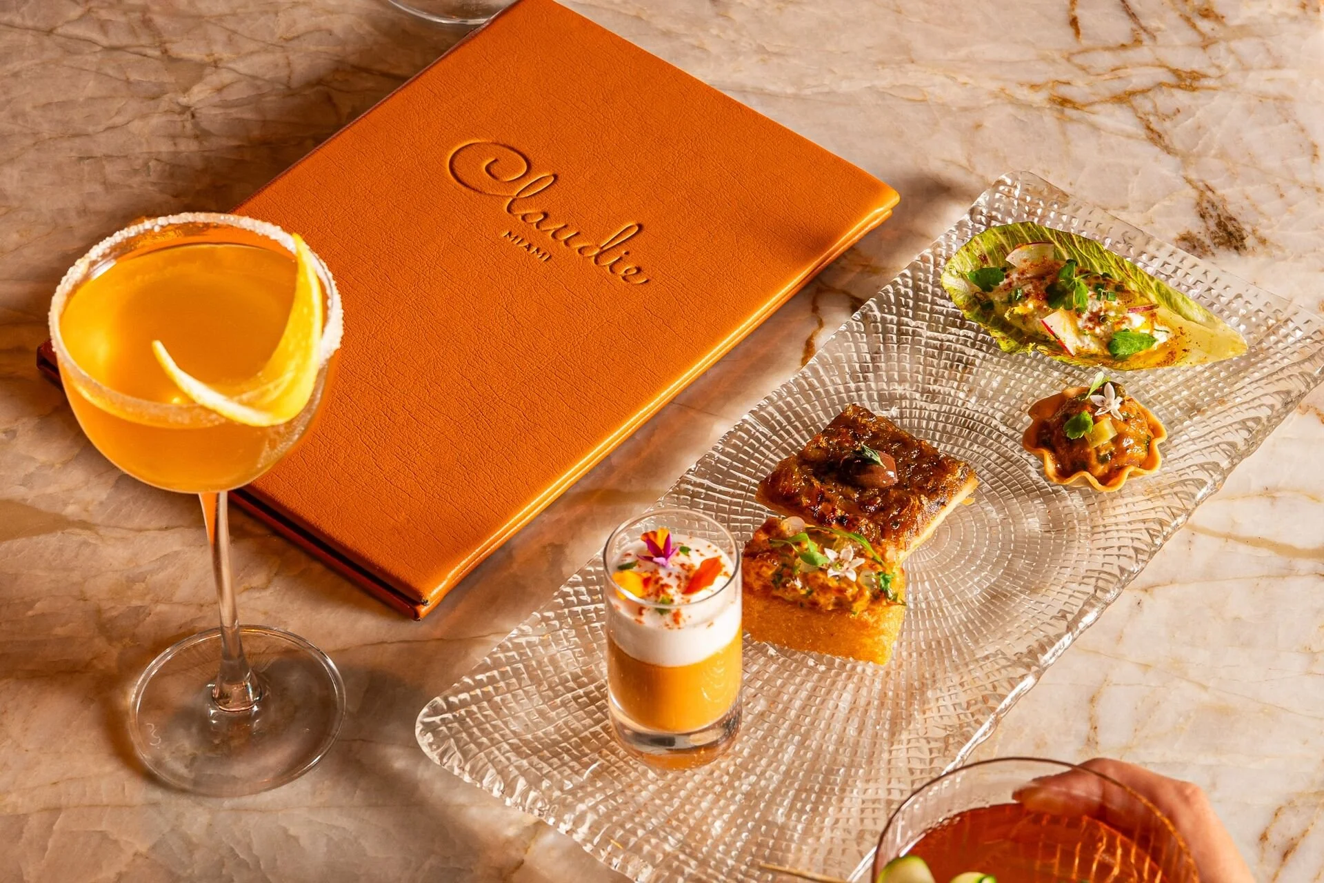 An elegant spread of appetizers and drinks on a marble table, including a cocktail with a lemon wedge, a layered shot with orange and cream, and a glass of sparkling wine, alongside a textured orange menu titled 'Claudine Miami' and a clear glass serving tray with assorted small bites and garnishes.