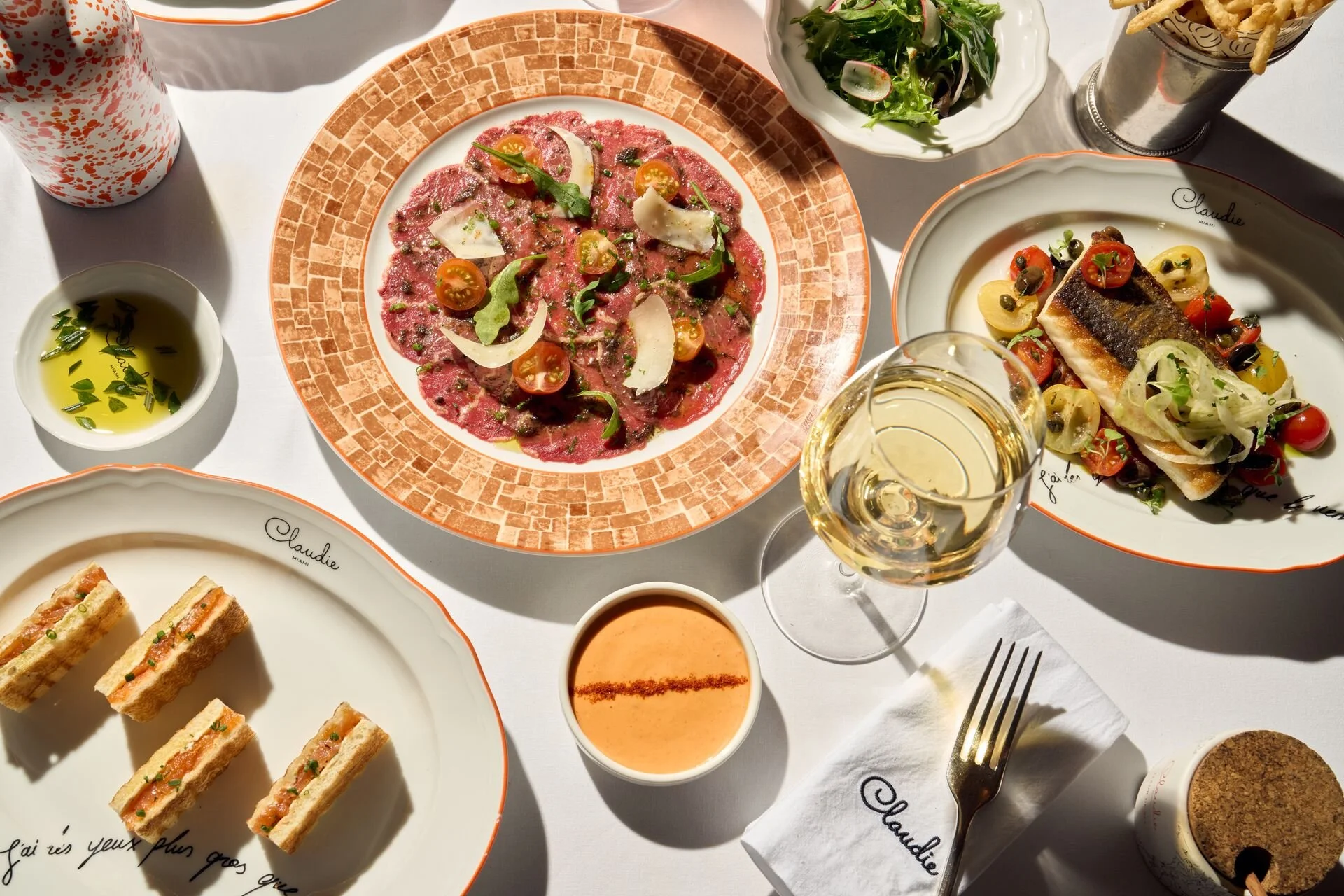 A table with various dishes including slices of beef carpaccio topped with cherry tomatoes, arugula, and shaved Parmesan, a plate of fish with cherry tomatoes and capers, a bowl of mixed greens, a small bowl of olive oil and herbs, a glass of white wine, and other smaller dishes, all set on a white tablecloth.