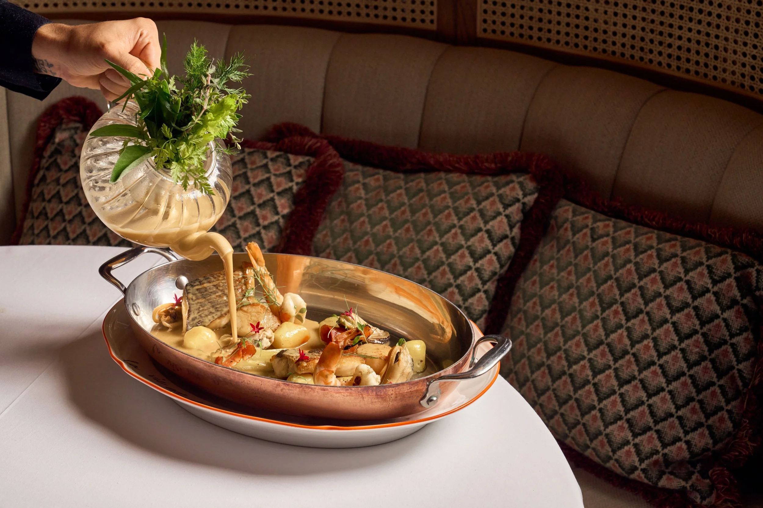 A hand placing fresh herbs into a copper serving dish with seafood and vegetables at a restaurant.