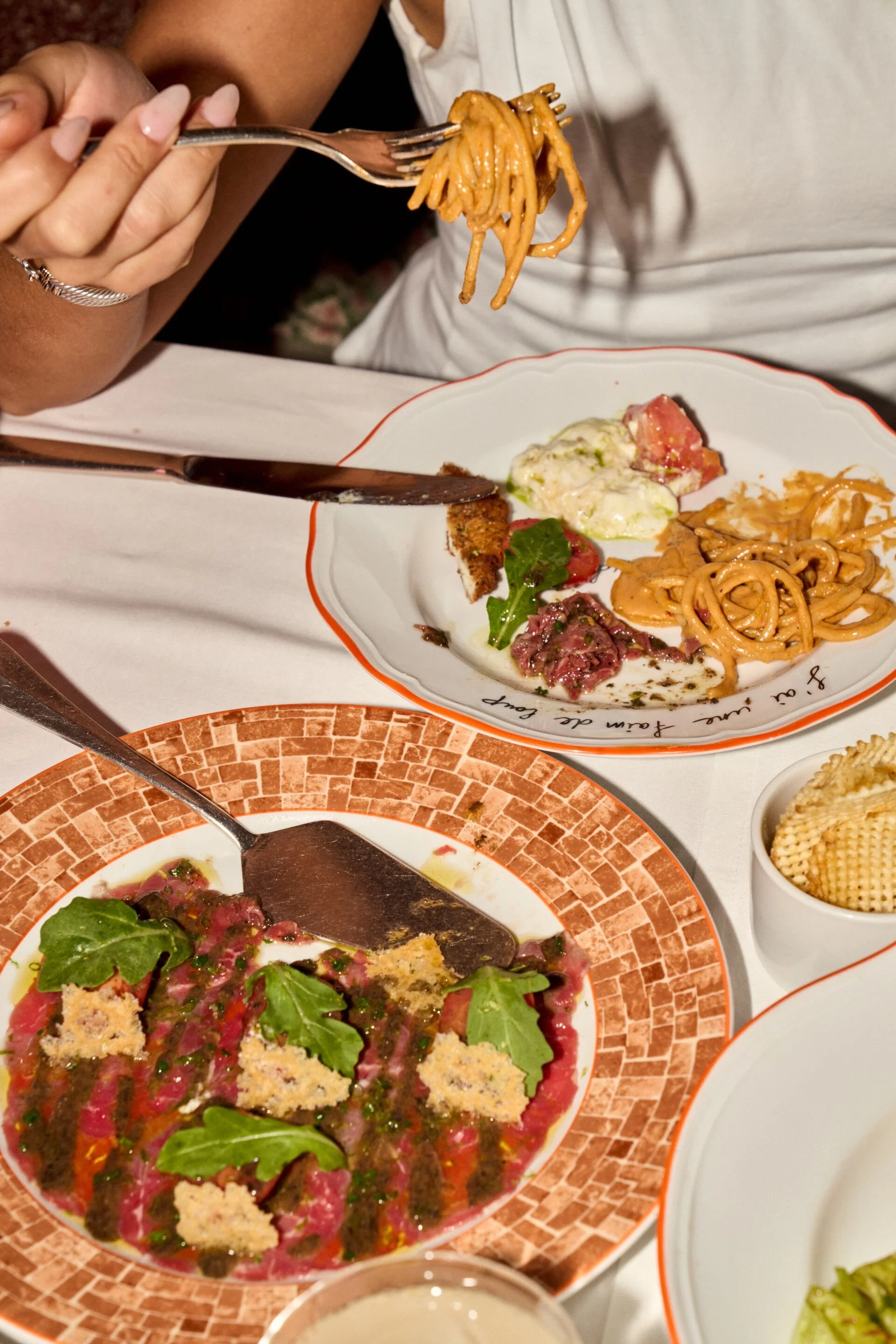 Close-up of a person holding a fork with spaghetti, with an appetizer and salad on a decorative plate on a white tablecloth.