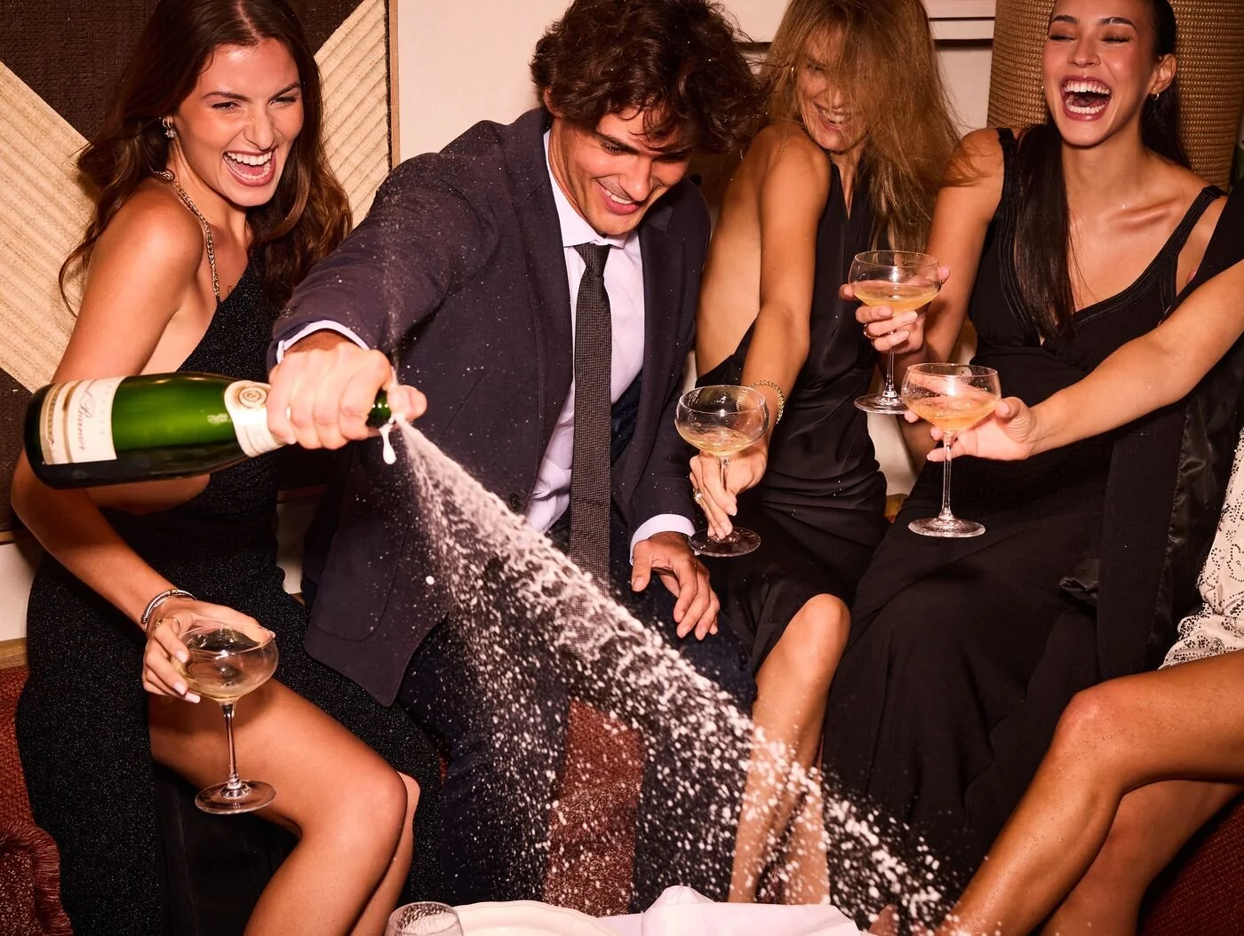 Group of friends celebrating with champagne, with one person spraying champagne, all are dressed in formal attire, smiling and laughing.