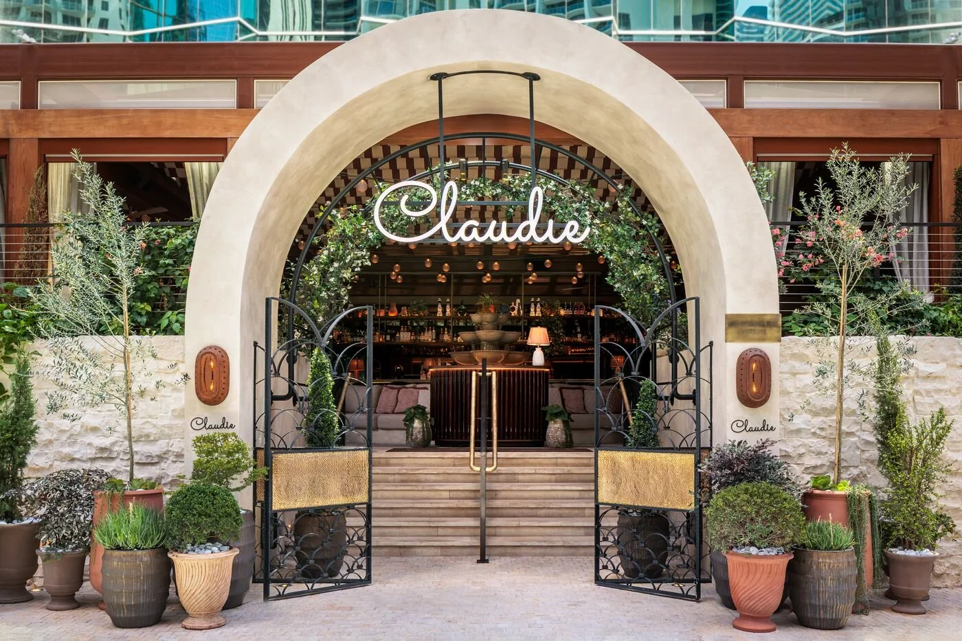 The entrance to a restaurant named Claudie with a modern arched design, black decorative gates, greenery, and potted plants outside, featuring warm lighting and an interior bar visible through glass.