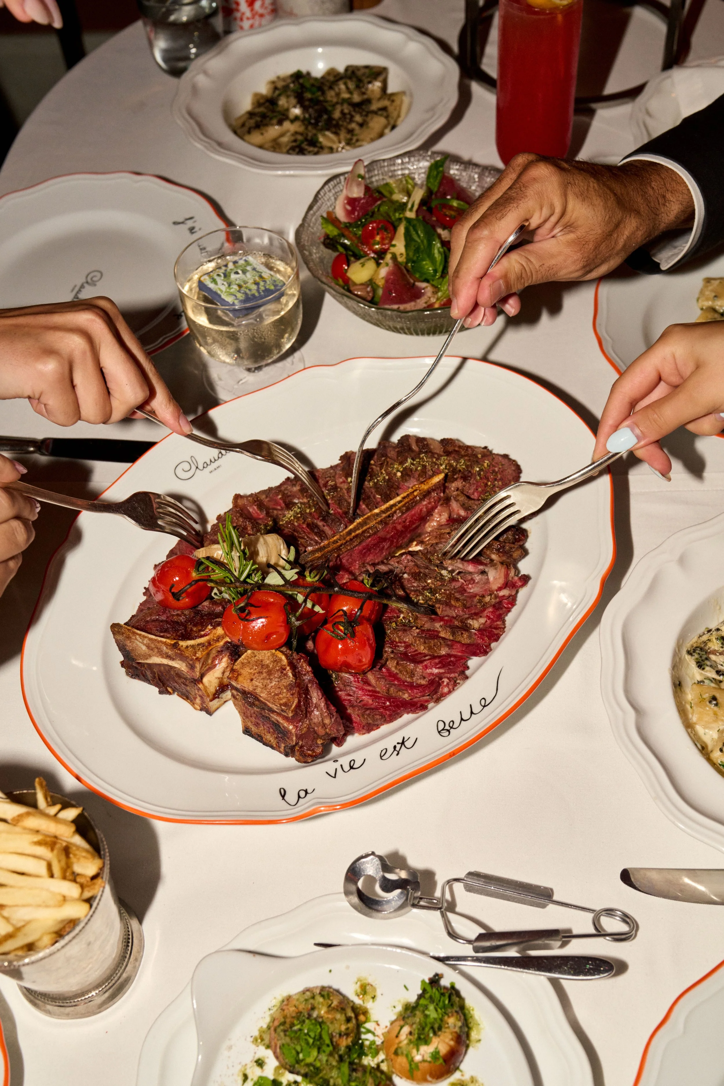 A dining table with plates of food, including a large roasted meat dish garnished with cherry tomatoes, herbs, and vegetables, a bowl of salad with tomatoes and greens, a plate of pasta, and other side dishes. People are using forks to serve themselves.