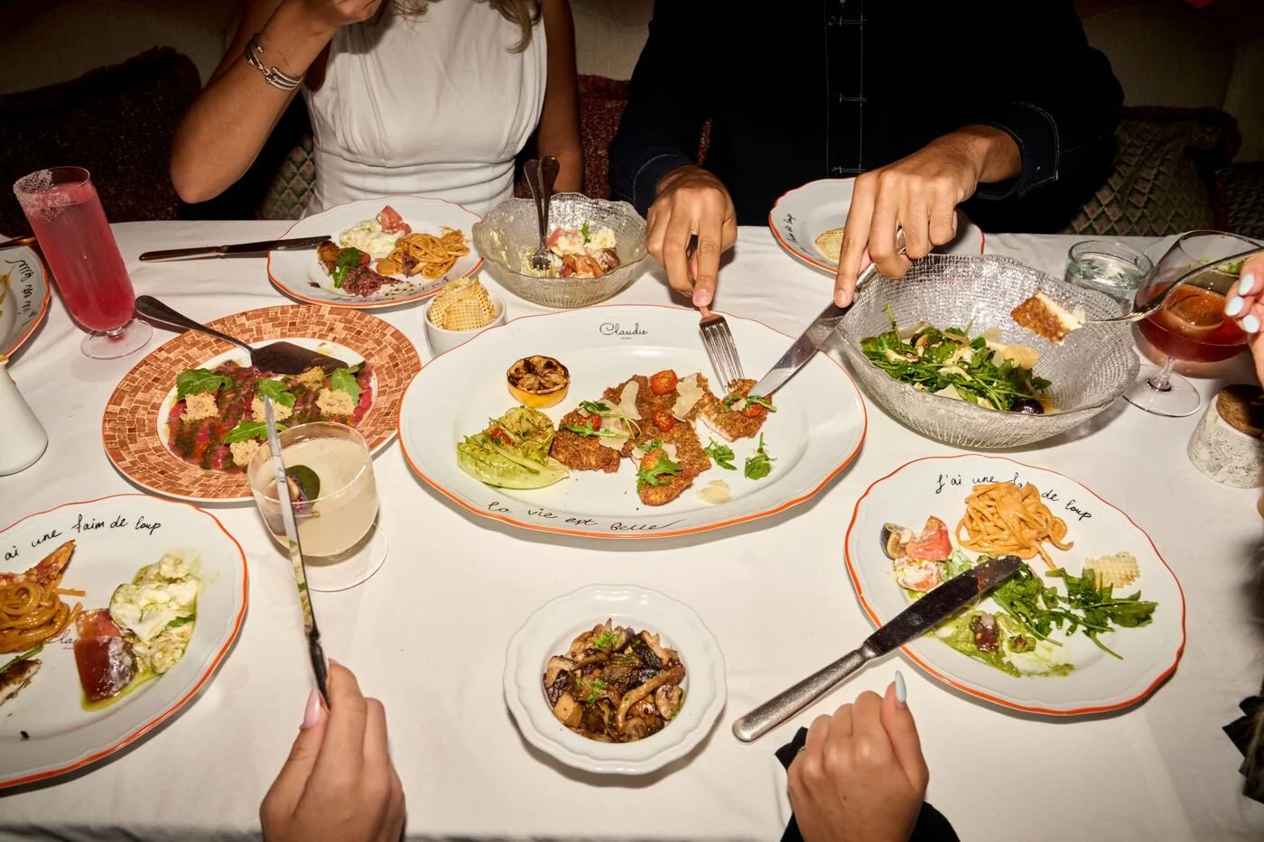 People gathered around a dinner table with various plates of food, including salads, pasta, and grilled dishes, sharing a meal.