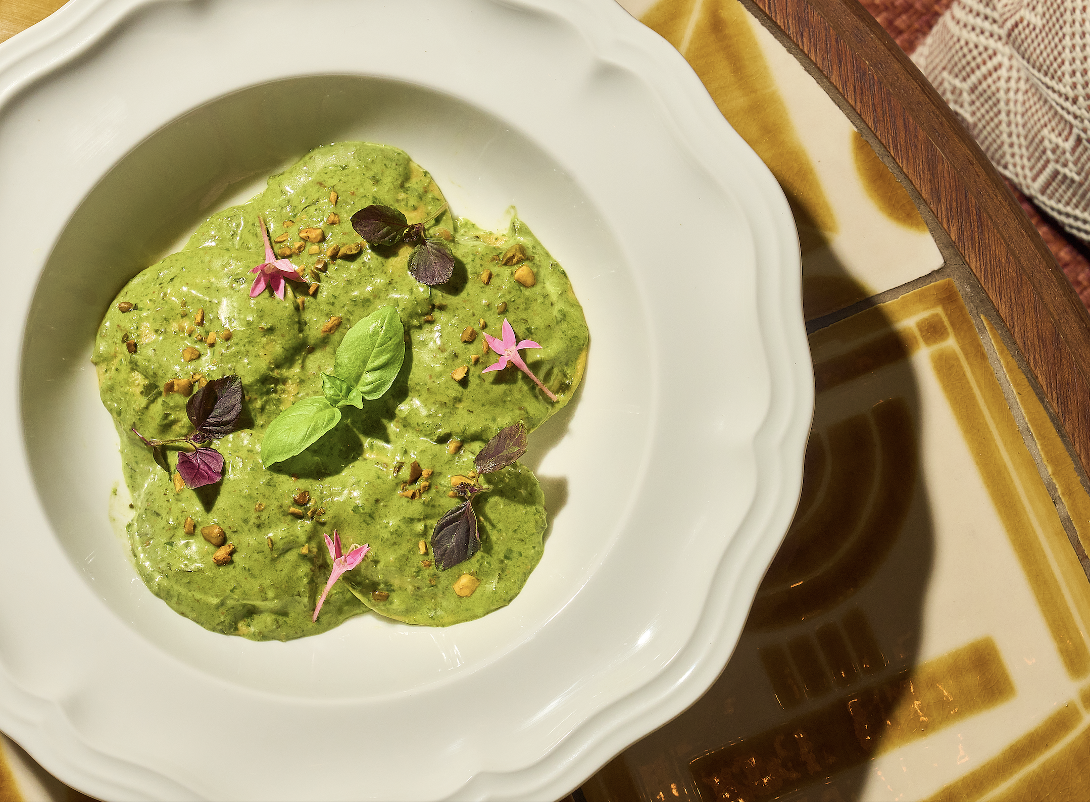 Close-up of a white bowl with green guacamole, garnished with chopped nuts, fresh basil, and pink edible flowers, placed on a wooden table.