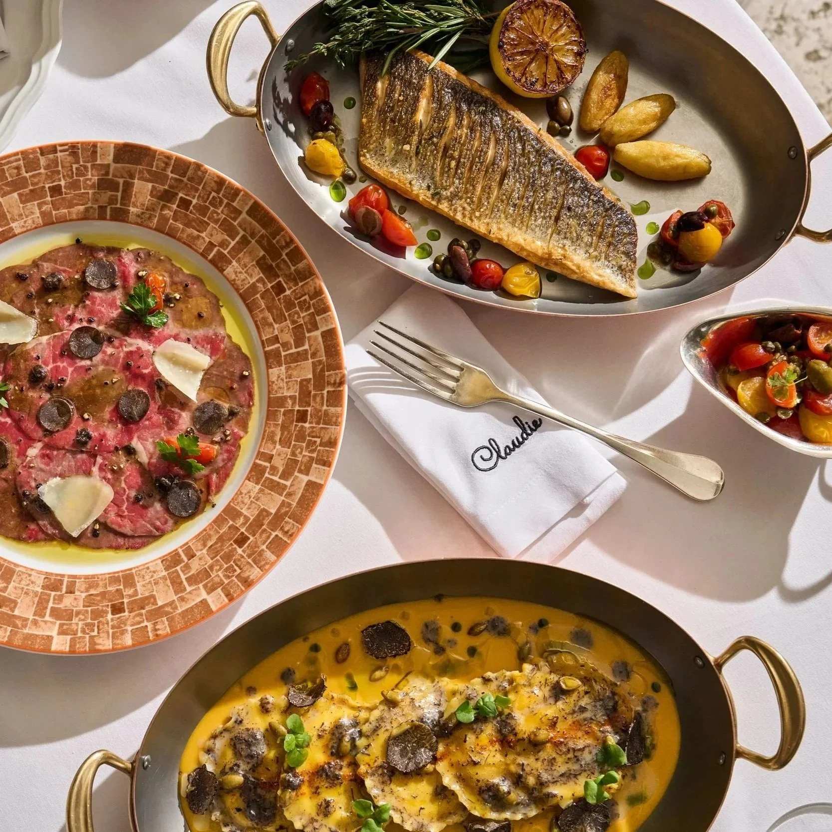 Assorted dishes including a grilled fish with roasted lemon and potatoes, a bowl of beef carpaccio with herbs and truffles, a vegetable medley salad, and a creamy yellow soup with mushrooms.