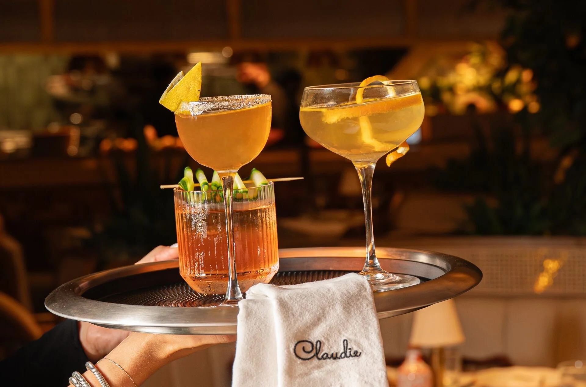 3 orange Cocktails from CLAUDIE's menu on a servers dish with a CLAUDIE napkin.
