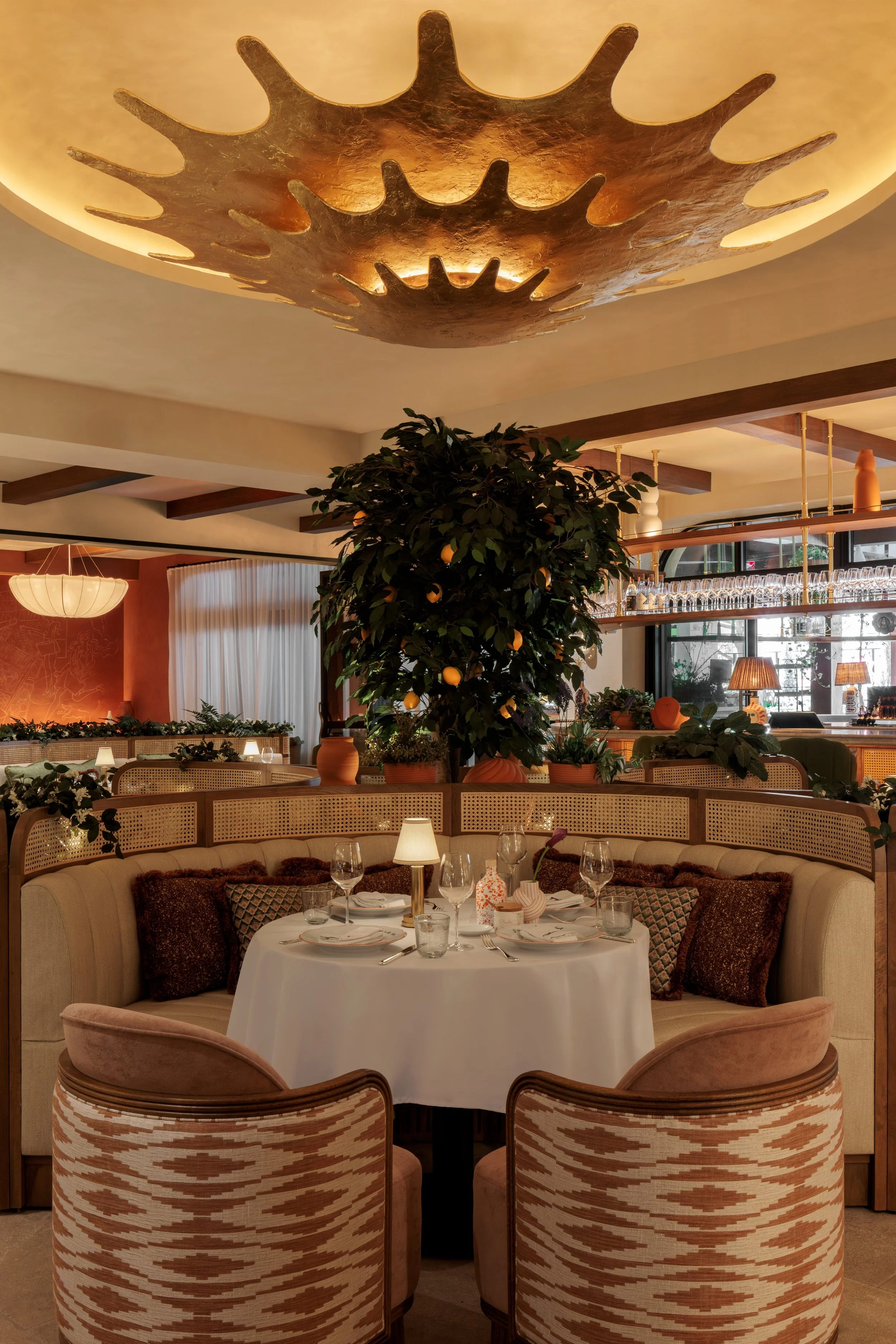 Inside a stylish restaurant with a round table set for dining, including wine glasses, utensils, and a candle; surrounded by cushioned chairs with patterned fabric and decorative pillows. A large greenery plant and a warm, modern lighting fixture are also visible.