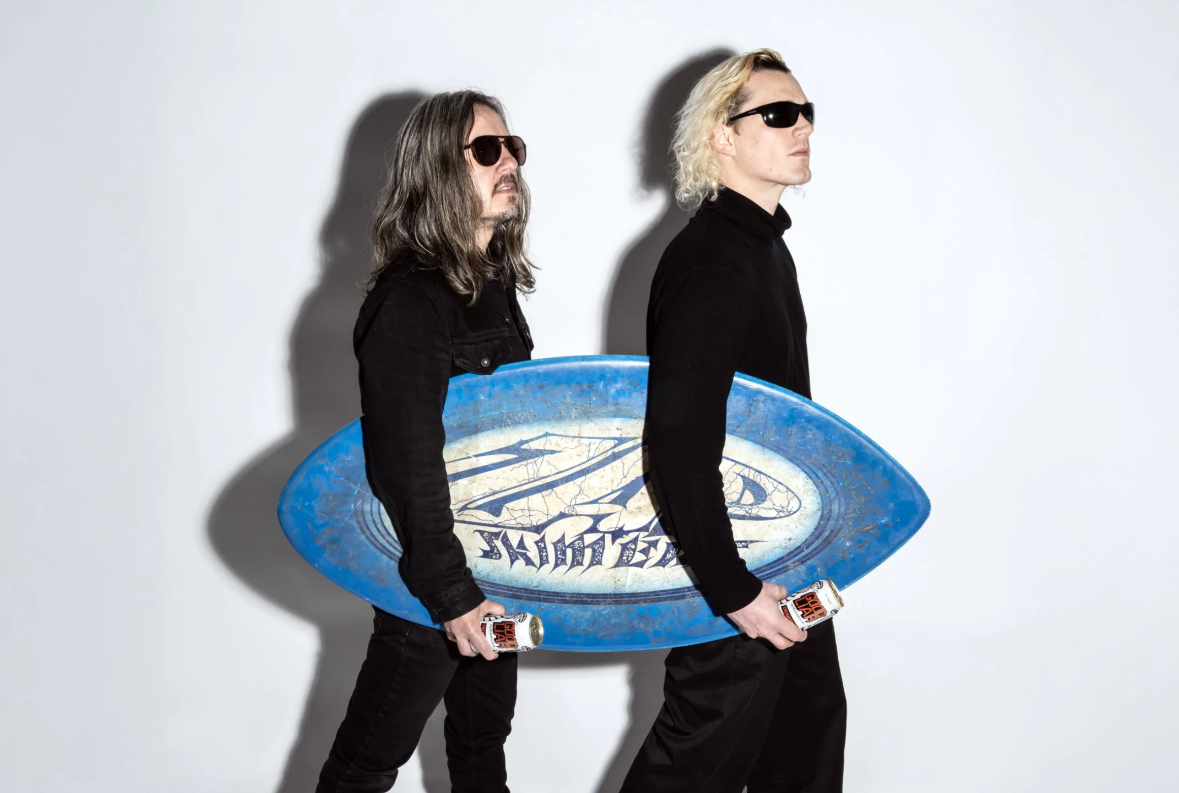 Two men wearing black clothing and sunglasses hold a blue surfboard with a logo, each holding a can of soda, standing against a white background.