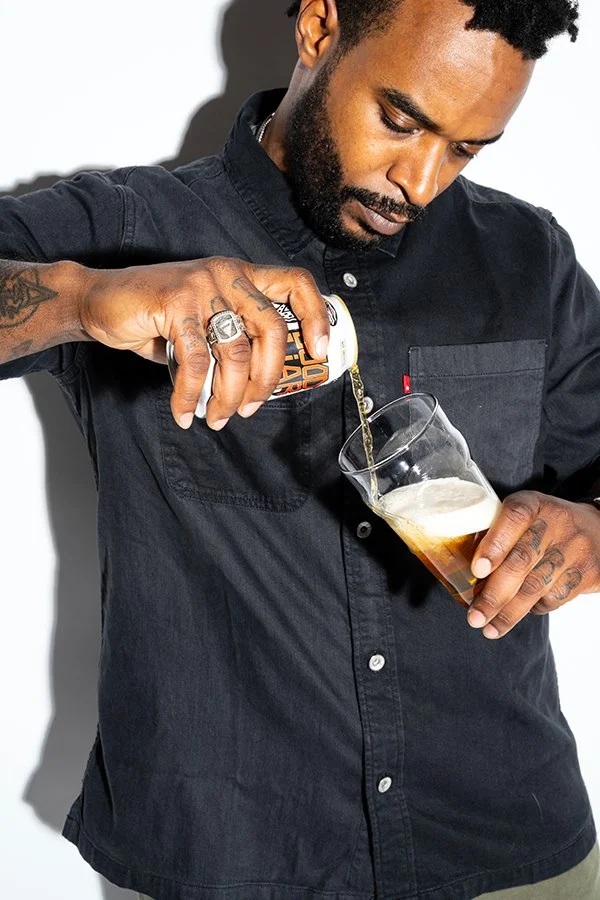 A man with tattoos and a black shirt is pouring beer from a can into a glass.