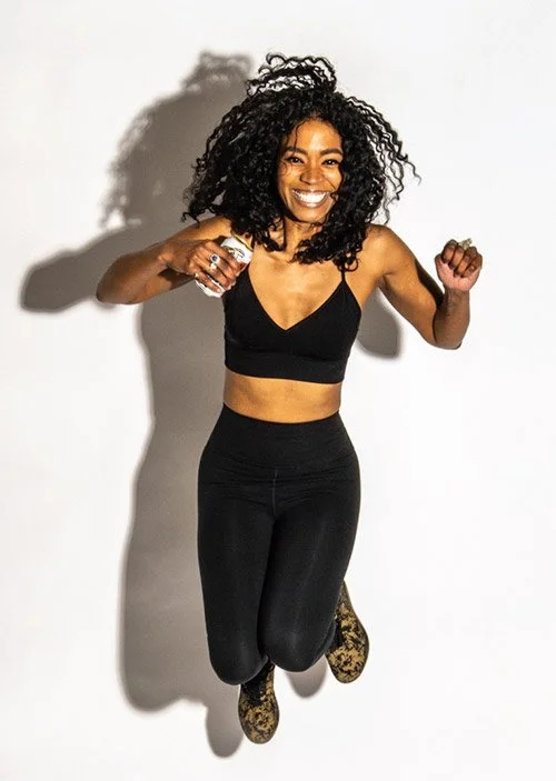 A woman with curly black hair jumping mid-air, smiling, wearing a black sports bra, black leggings, and patterned shoes, with a shadow cast on a white background.