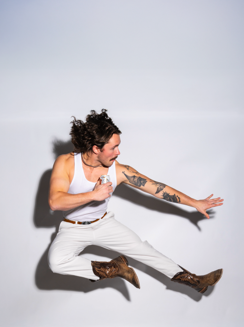 A man with curly hair and tattoos wearing a white tank top and white pants, jumping mid-air against a plain white background while holding a can of beer.