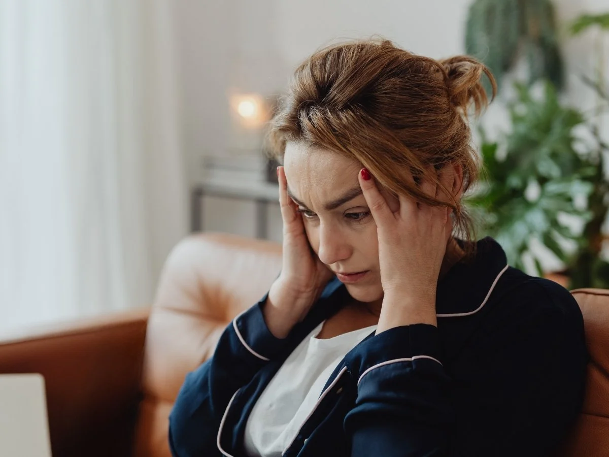 A woman sits on a couch pressing her fingers to her temples, looking down with a worried expression | Blue Lotus Telepsychiatry