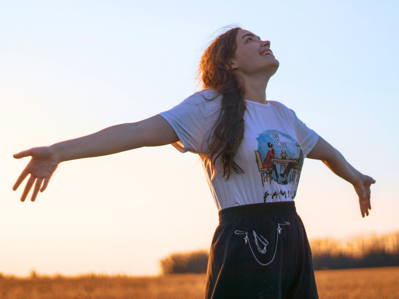 A woman stands in a field with her arms outstretched, smiling and looking up as the sun sets behind her | Blue Lotus Telepsychiatry