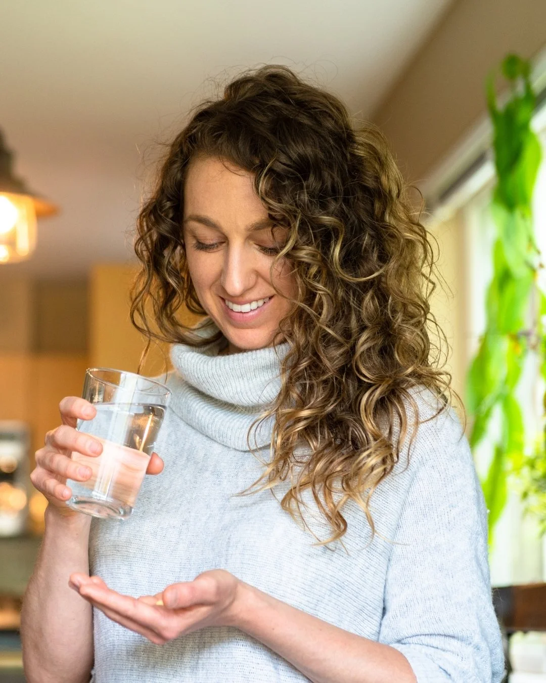 A woman holds a glass of water and a pill in her hand while smiling in a cozy home setting | Blue Lotus Telepsychiatry Services, Inc.