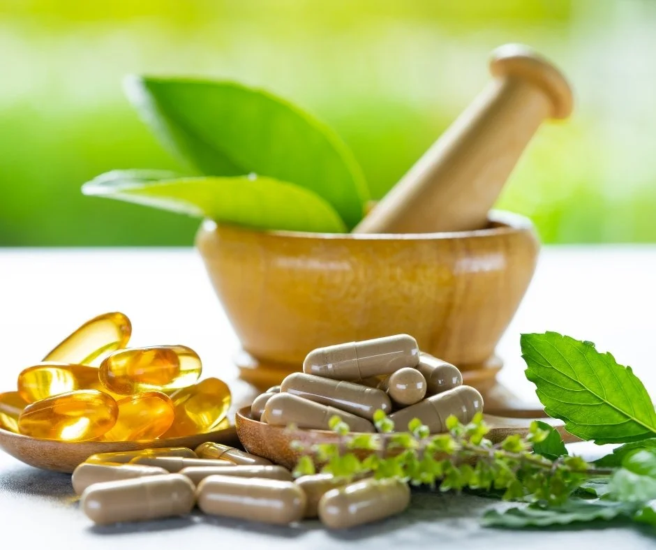 Herbal capsules and supplements sit beside a wooden mortar and pestle with fresh green leaves | Blue Lotus Telepsychiatry Services, Inc.