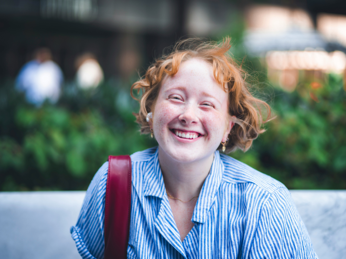 A woman with short, wavy red hair and a blue striped shirt smiling broadly outdoors while wearing a shoulder bag | Blue Lotus Telepsychiatry